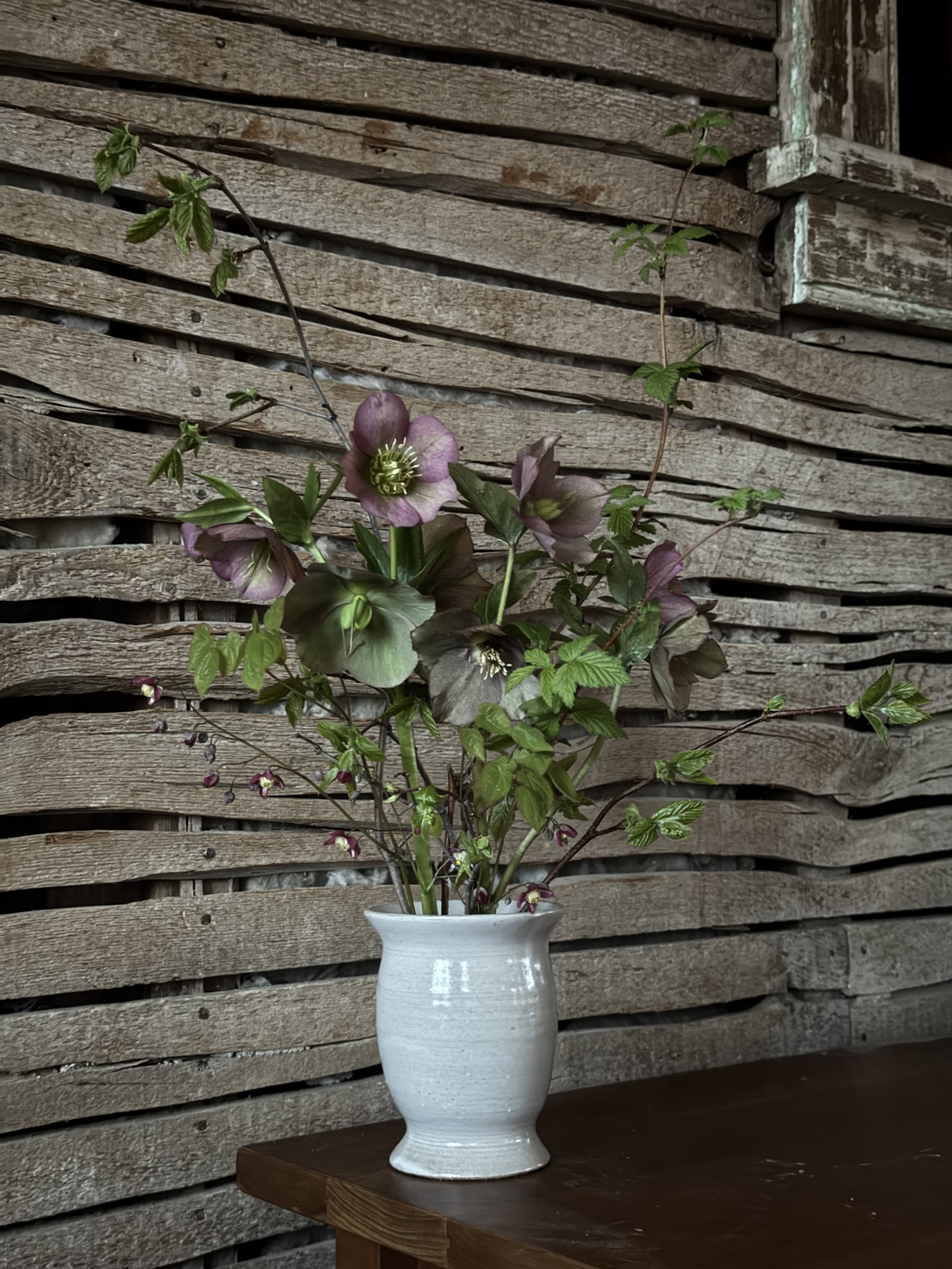 Vase with purple and white flowers and green foliage, placed on a wooden table against a background of weathered wooden planks.