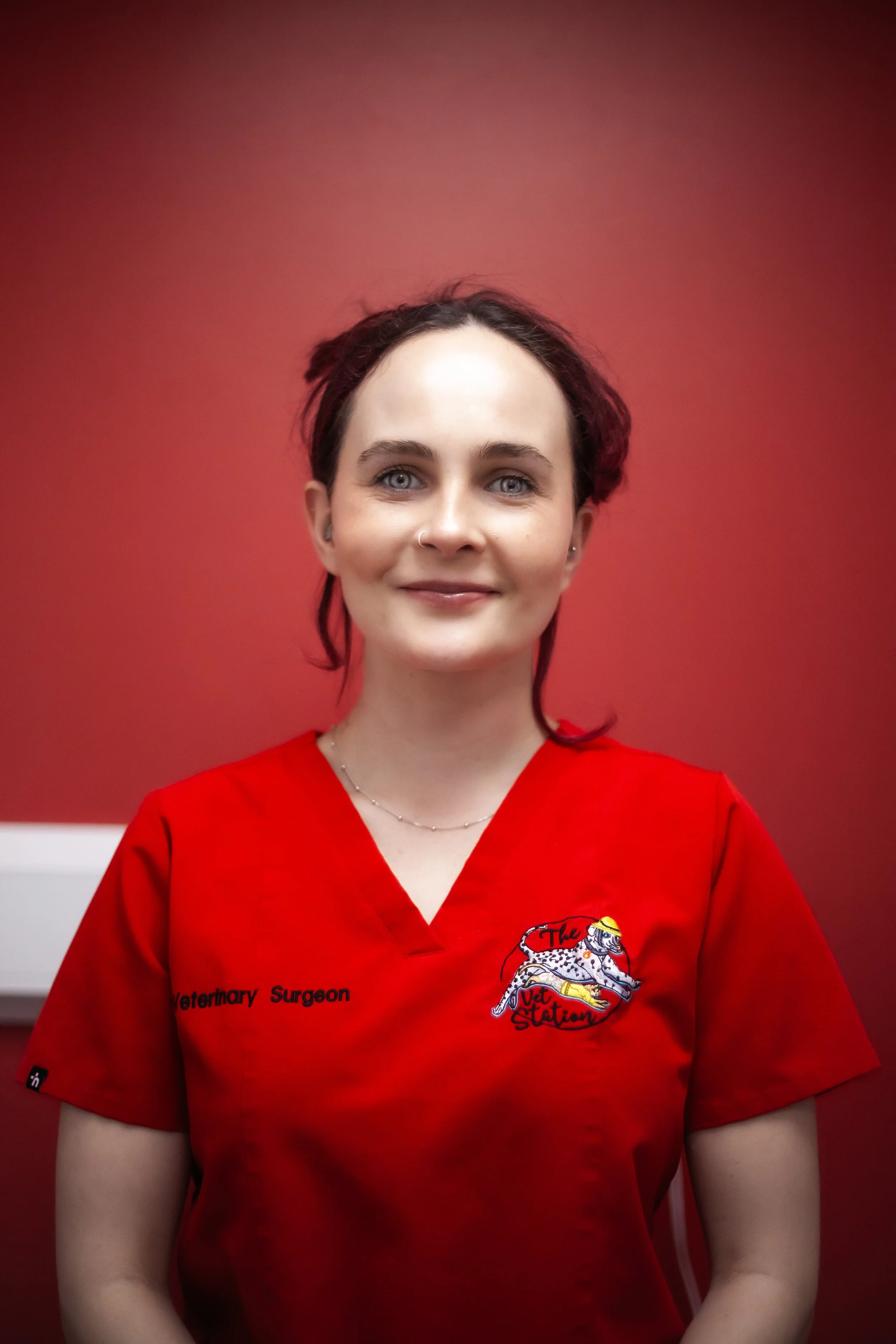 A woman with dark, wavy hair and a nose piercing, wearing a red veterinary surgical scrubs top with a logo and text, standing against a white brick wall.