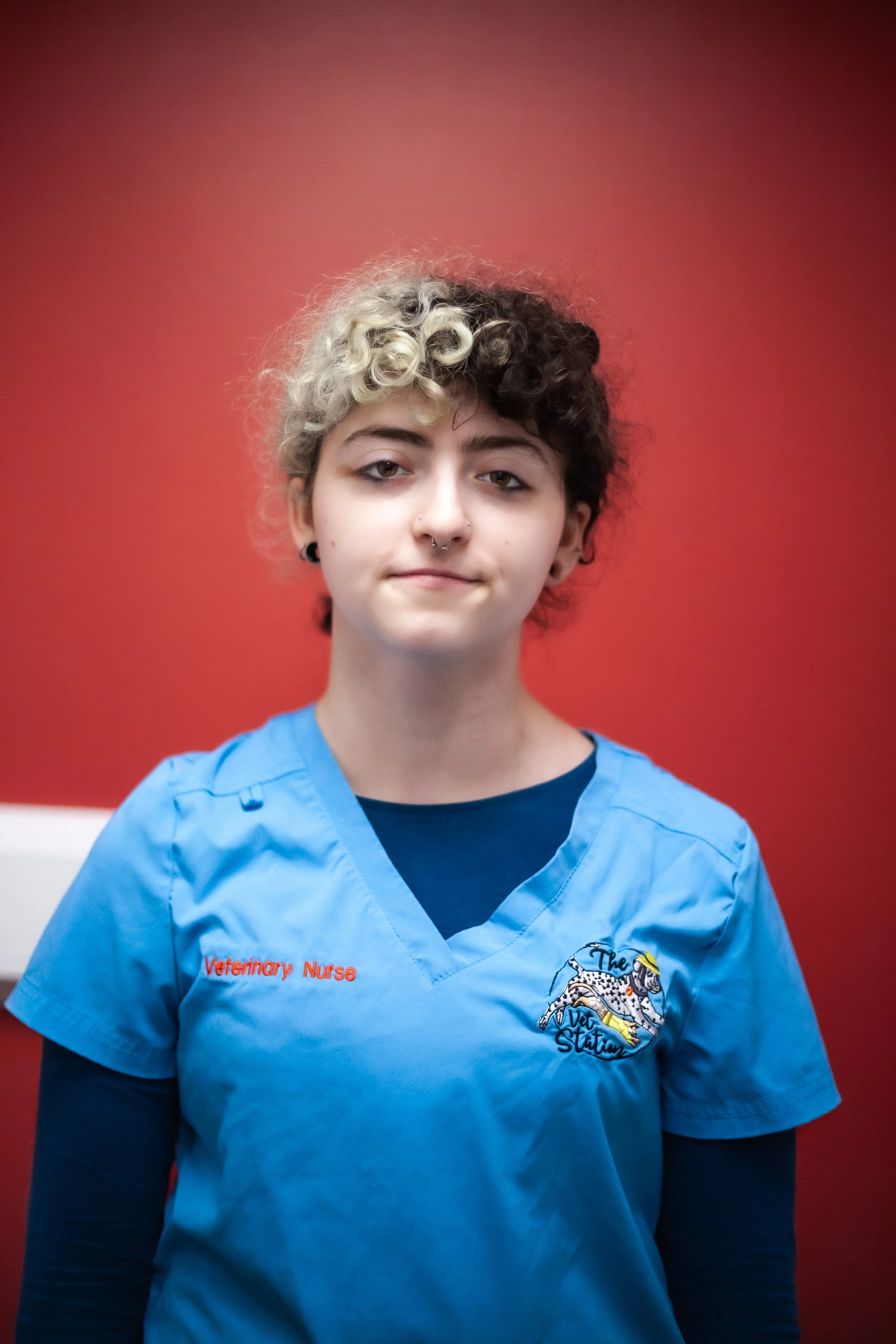 A young woman with curly hair, wearing a maroon veterinary nurse uniform with a name tag that reads 'Brenna, Student Veterinary Nurse'. She is smiling and standing in front of a white wall.