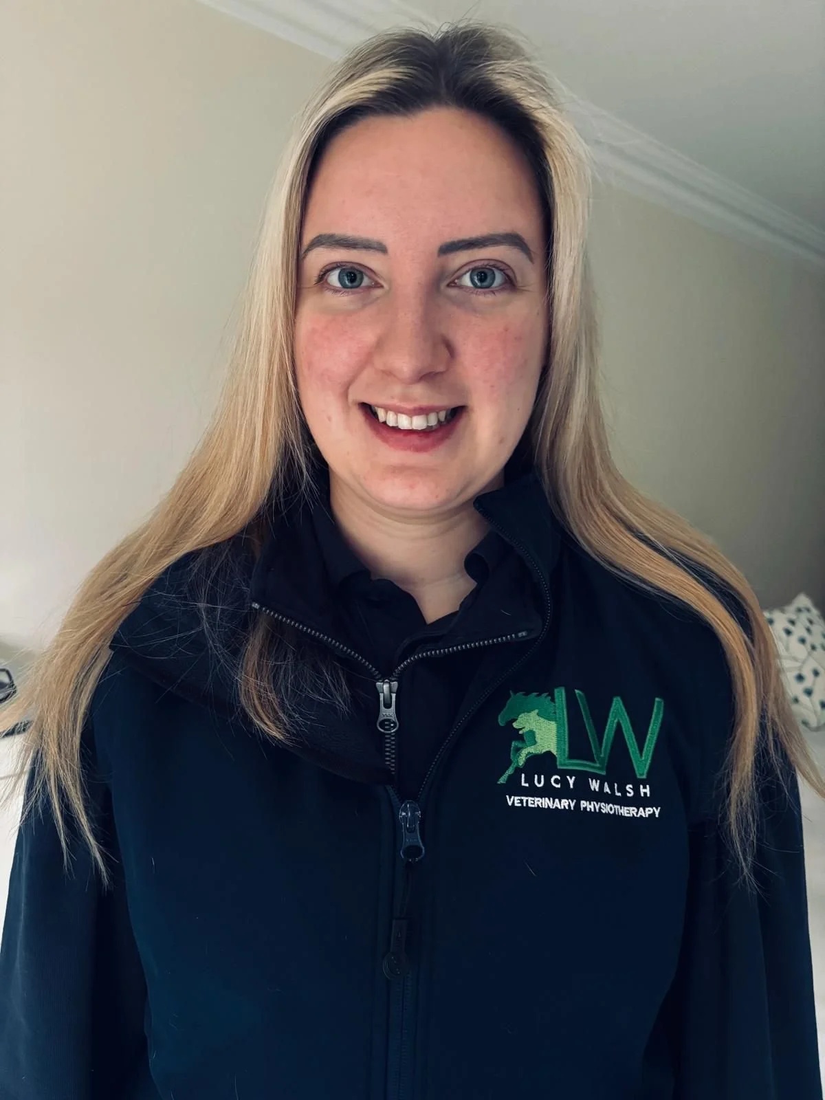 A woman with long blonde hair and blue eyes smiling, wearing a black jacket with a logo that says 'Lucy Walsh Veterinary Physiotherapy'.
