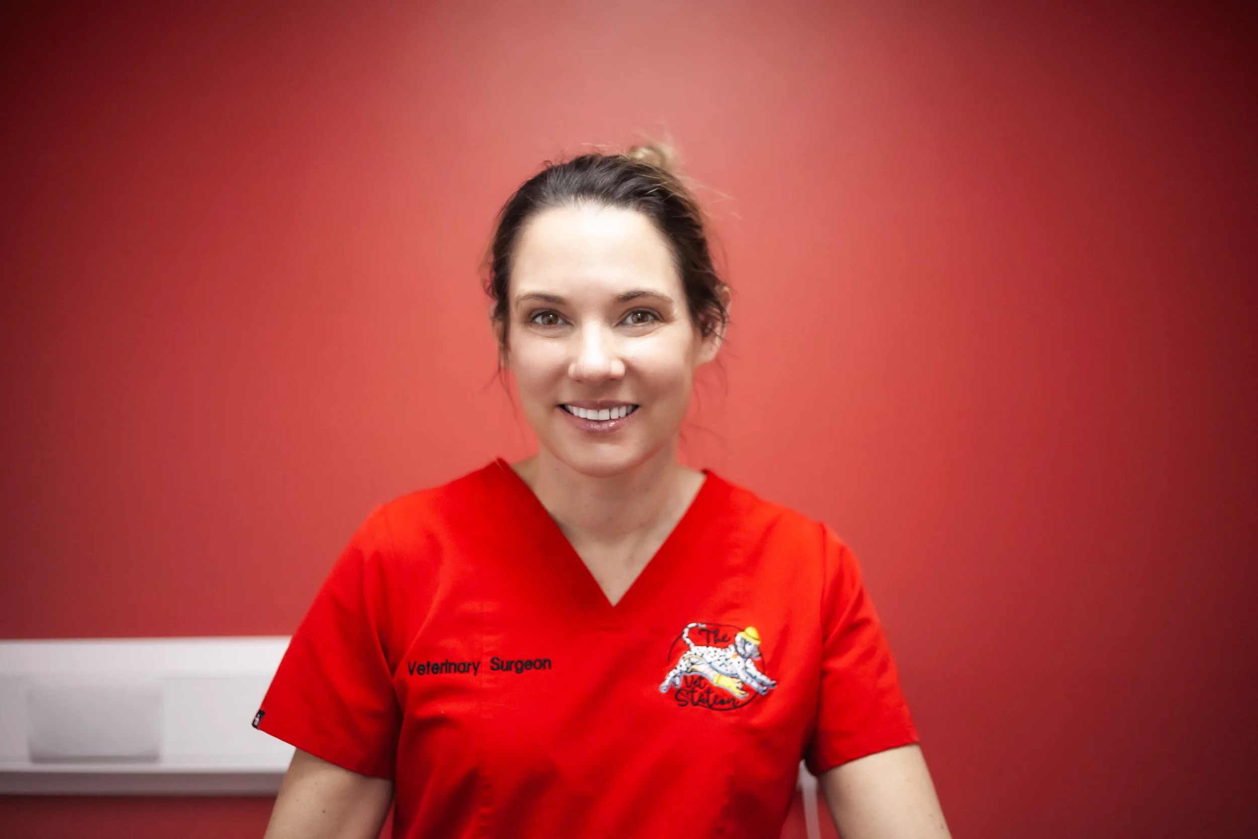 A woman in a red veterinary scrubs top smiling, with a red background and a white examination table behind her.
