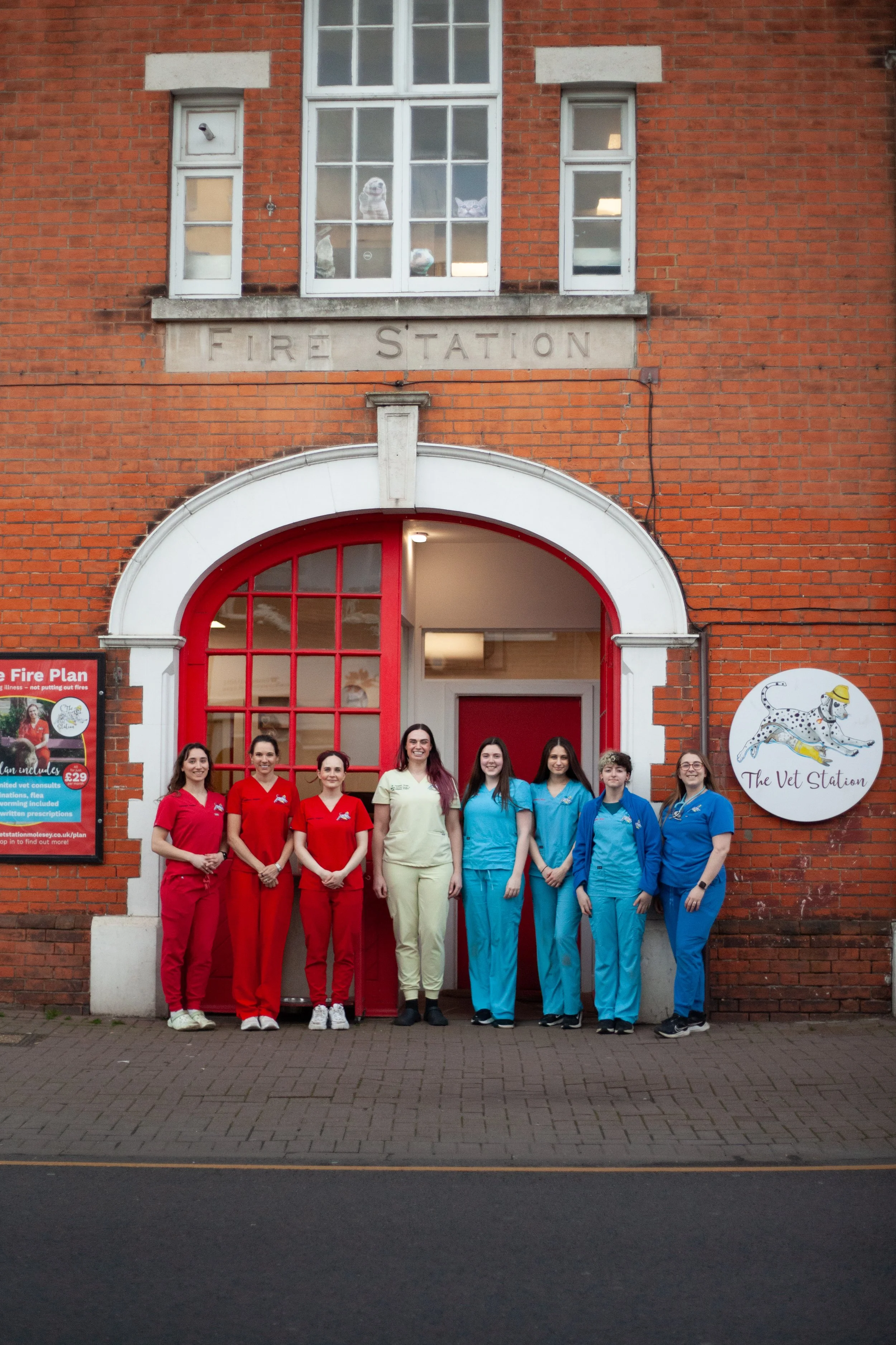Group of seven people, some in red and some in blue scrubs, standing outside a brick fire station building with a red door and a white sign that says "The Vet Station."