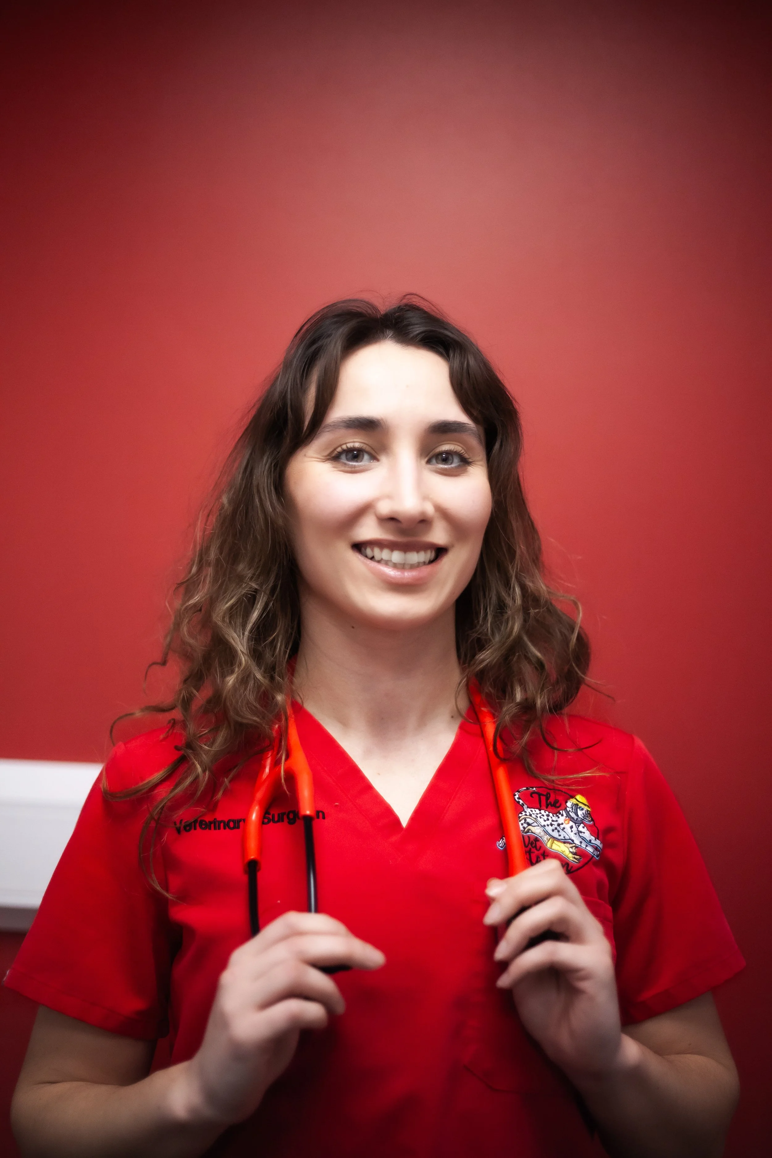 A young woman with long, wavy brown hair wearing a red jacket and holding a yellow stethoscope in front of a white brick wall.