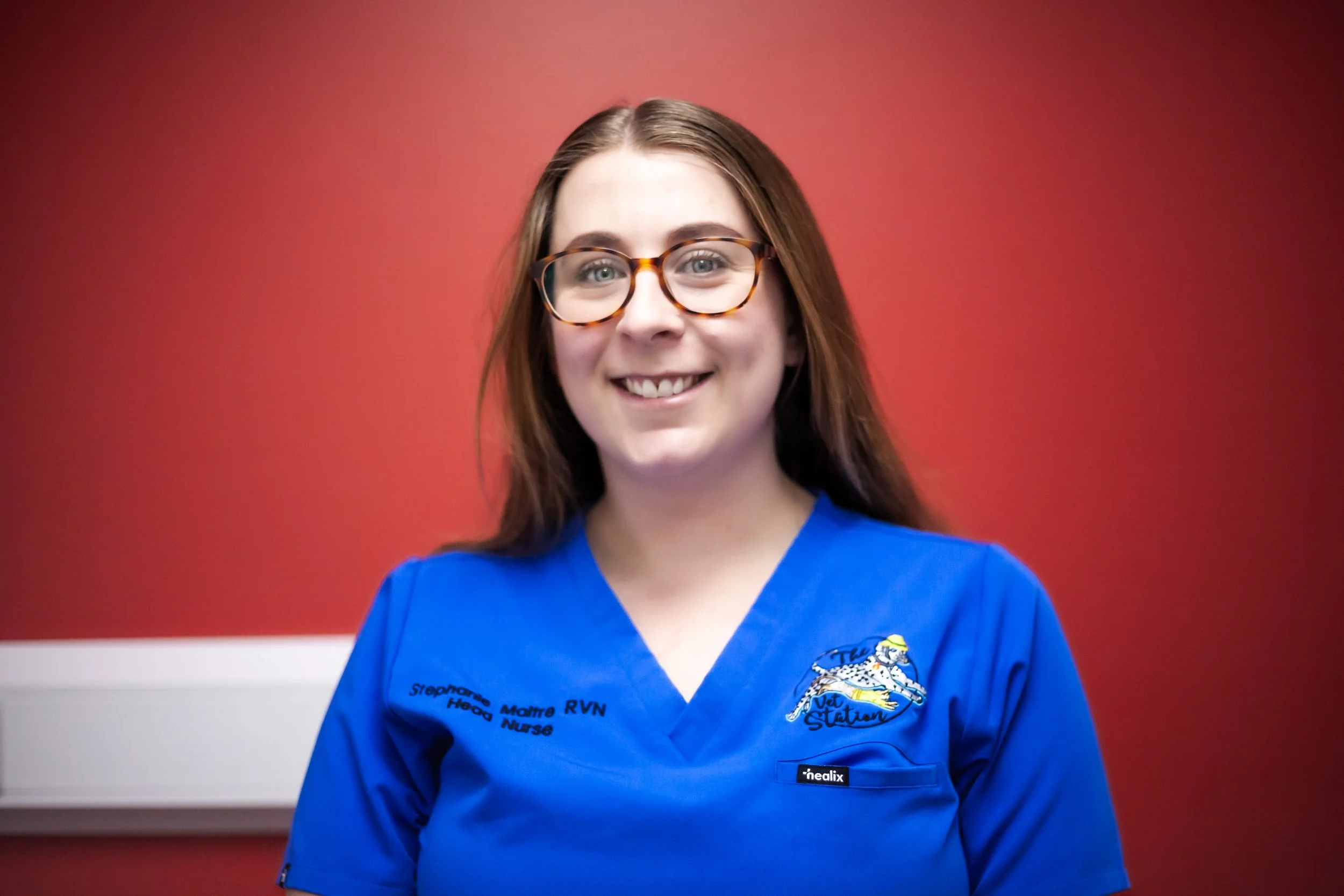 A woman with long brown hair wearing glasses and a blue medical uniform standing against a white wall.