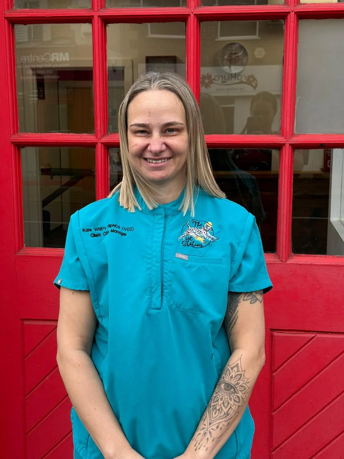 Smiling woman in a maroon uniform with a badge that reads 'Kate' standing against a white brick wall.