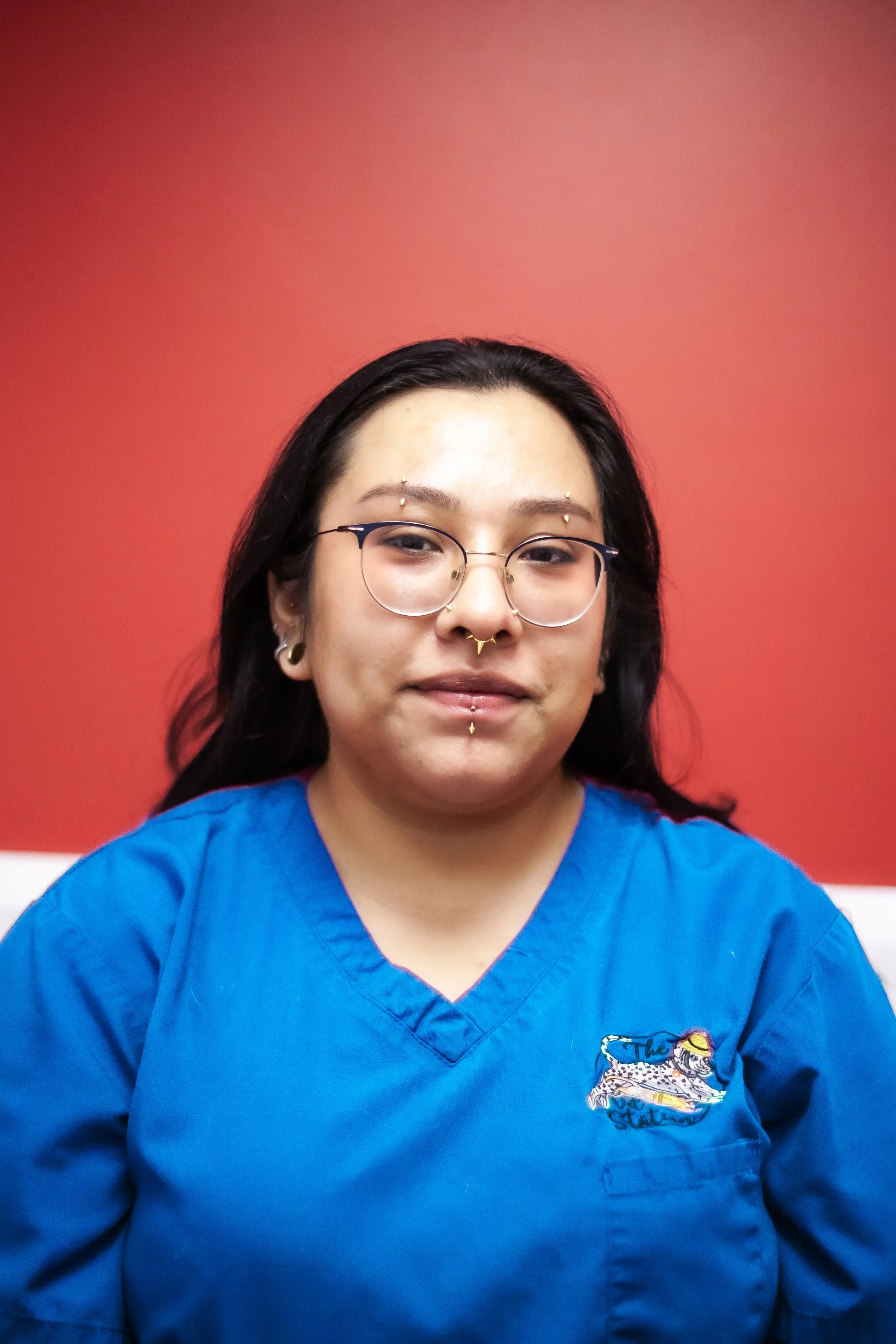 Portrait of a woman with glasses, piercings, and tattoos, wearing a blue scrubs against a red wall.