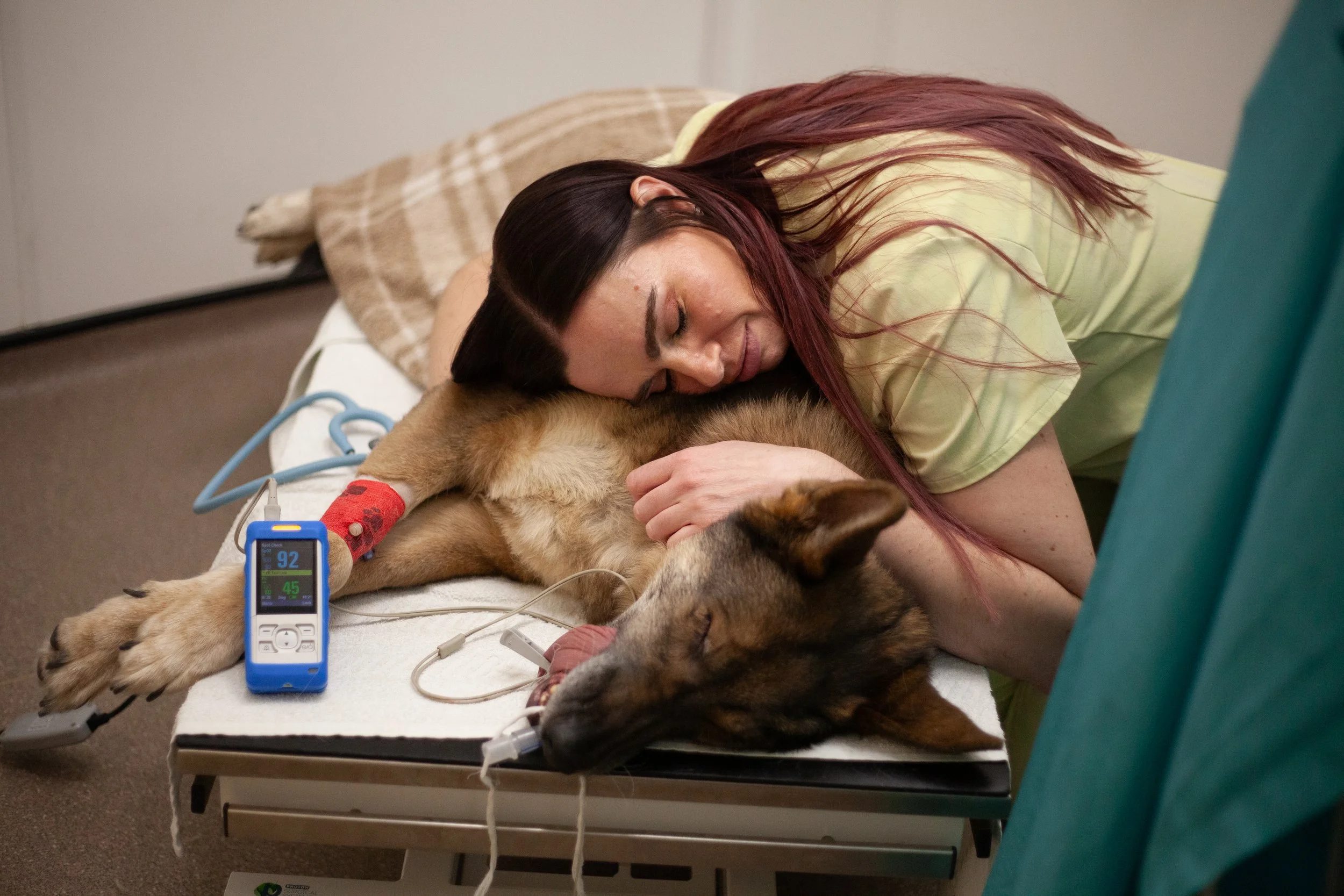 A woman with long red hair hugging a dog on a veterinary examination table. The dog has a medical bandage with red spots on its paw, a heart monitor, and is connected to medical equipment. The woman and dog appear to be resting peacefully.