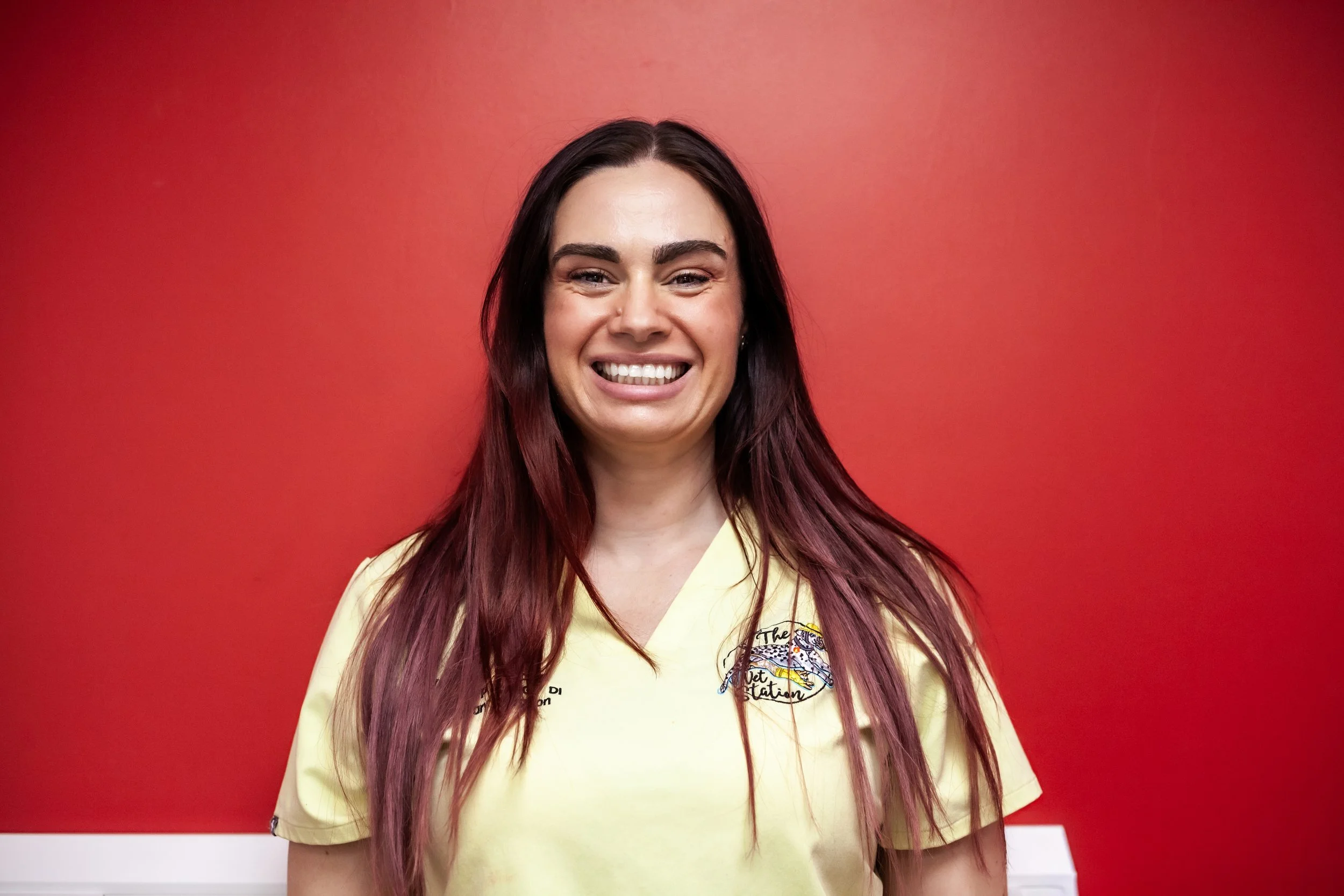 A woman smiling, wearing a red uniform with a vet logo, standing against a white brick wall.
