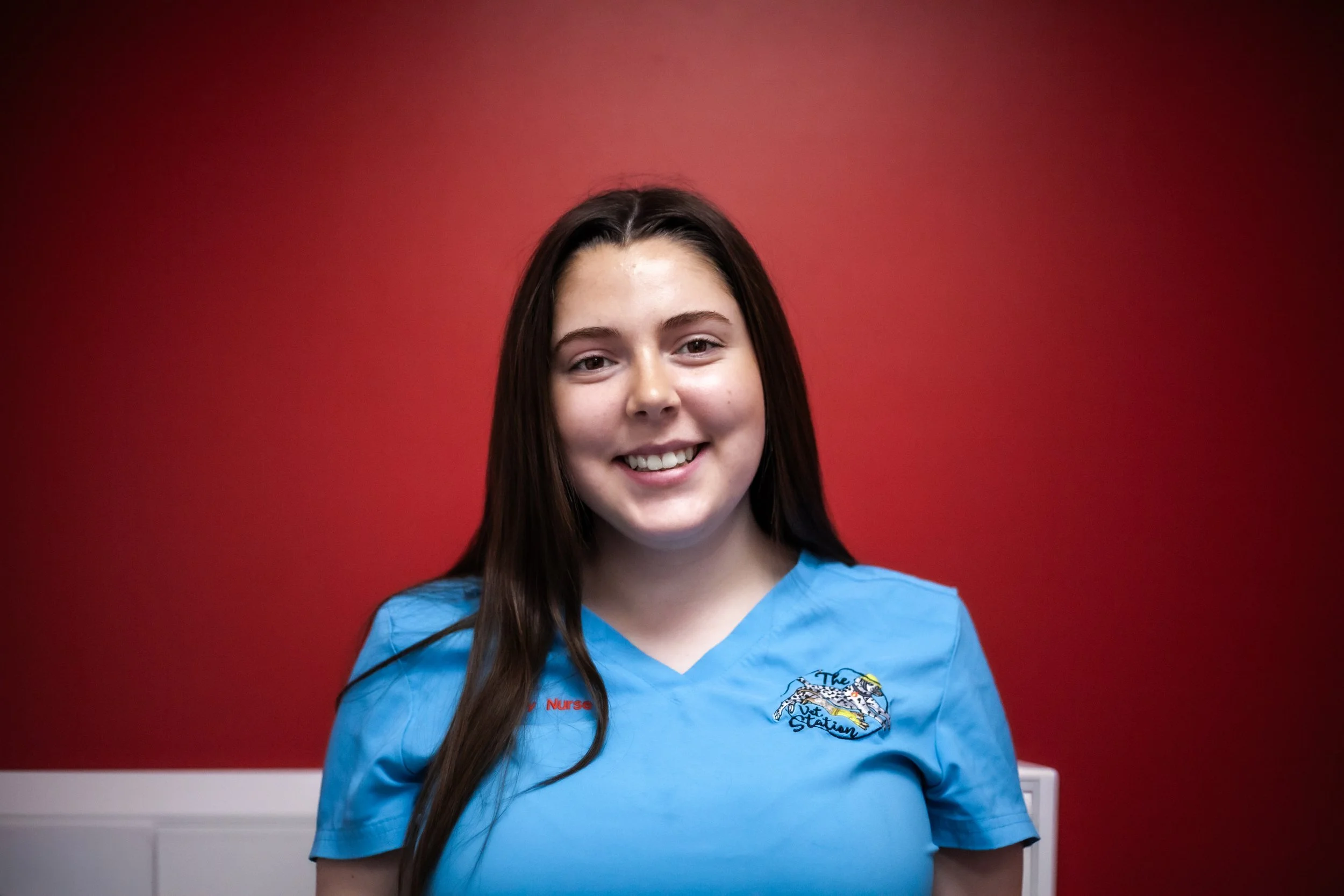 A young woman with dark hair in a braid, wearing blue scrubs with 'Veterinary Nurse' embroidered on the left side, smiling and standing against a white brick wall.