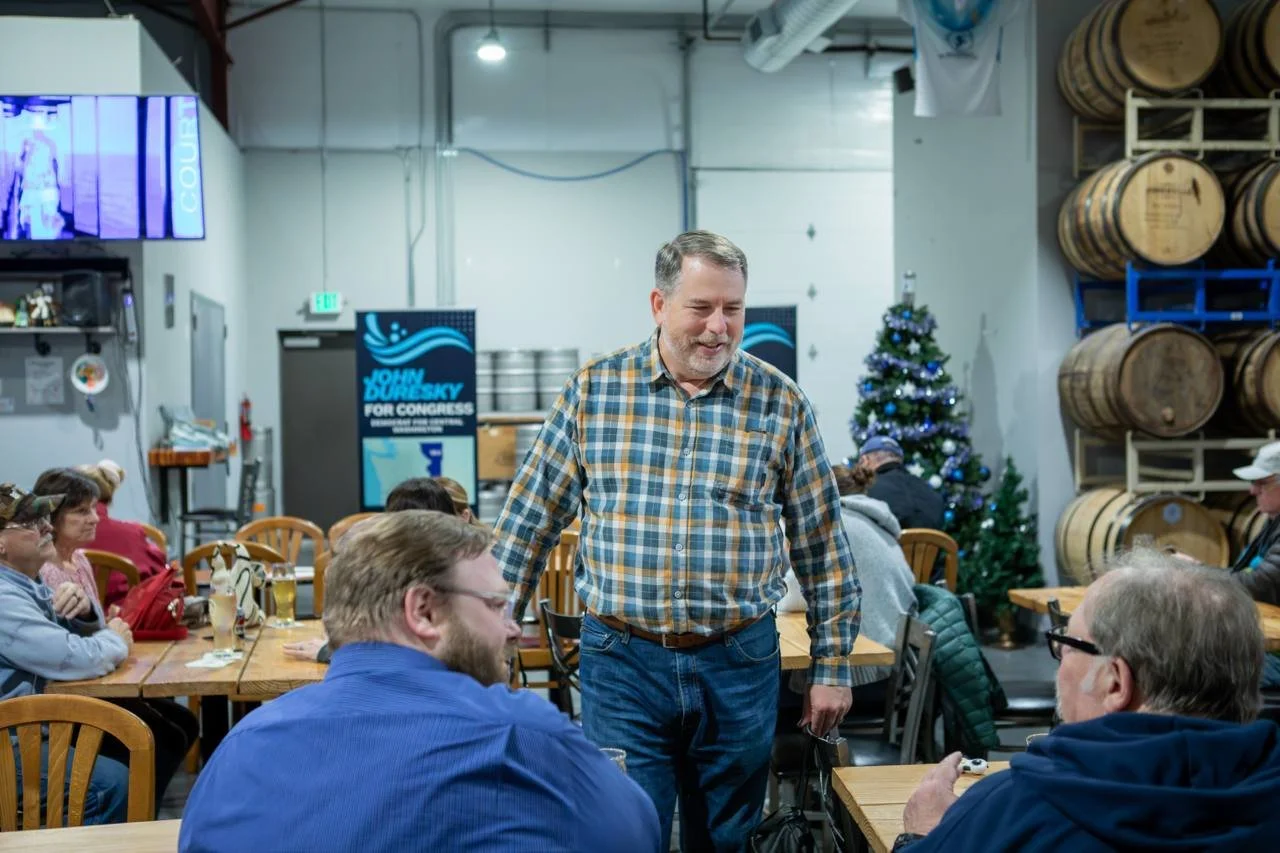 A man in a plaid shirt stands and chats with seated people at a gathering in a brewery, with a decorated Christmas tree and barrels in the background.
