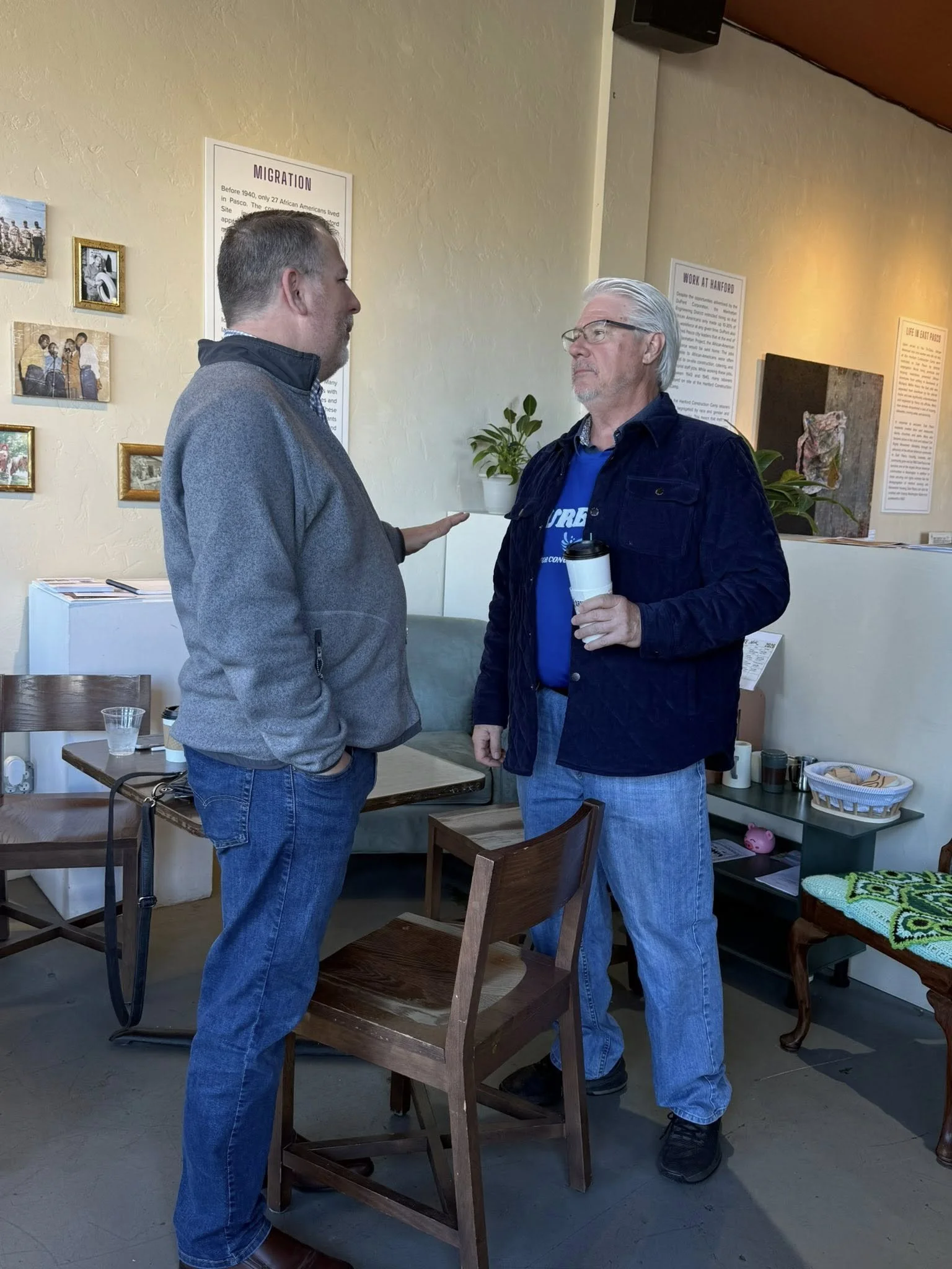 Two men in casual clothing stand and talk indoors, one holding a coffee mug, with relaxed postures and serious expressions.