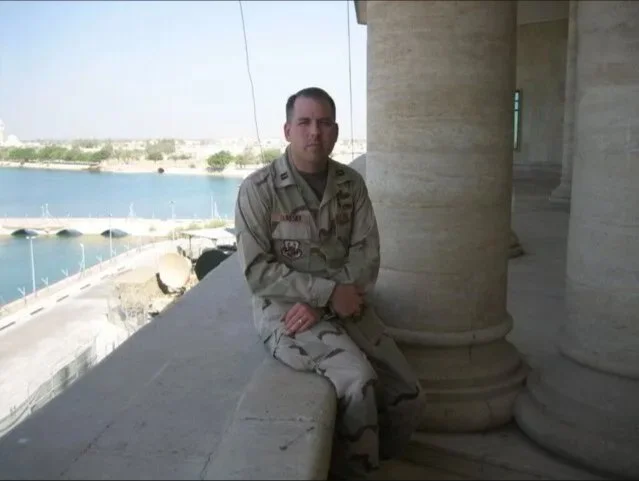 A man in military uniform sitting on a ledge outside a building with large stone columns, overlooking a body of water with boats and a distant shoreline.