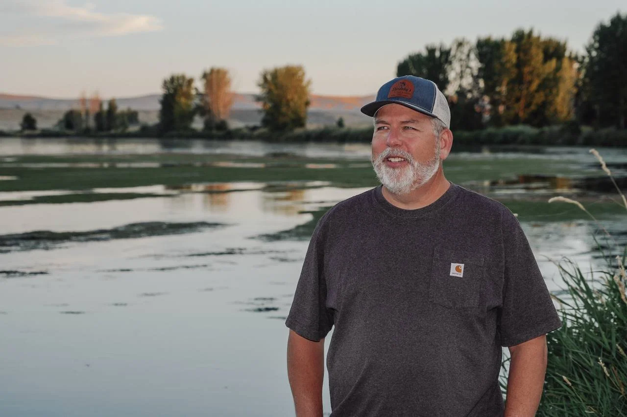 John Duresky stands outdoors near the Columbia River with trees and hills in the background during sunset.
