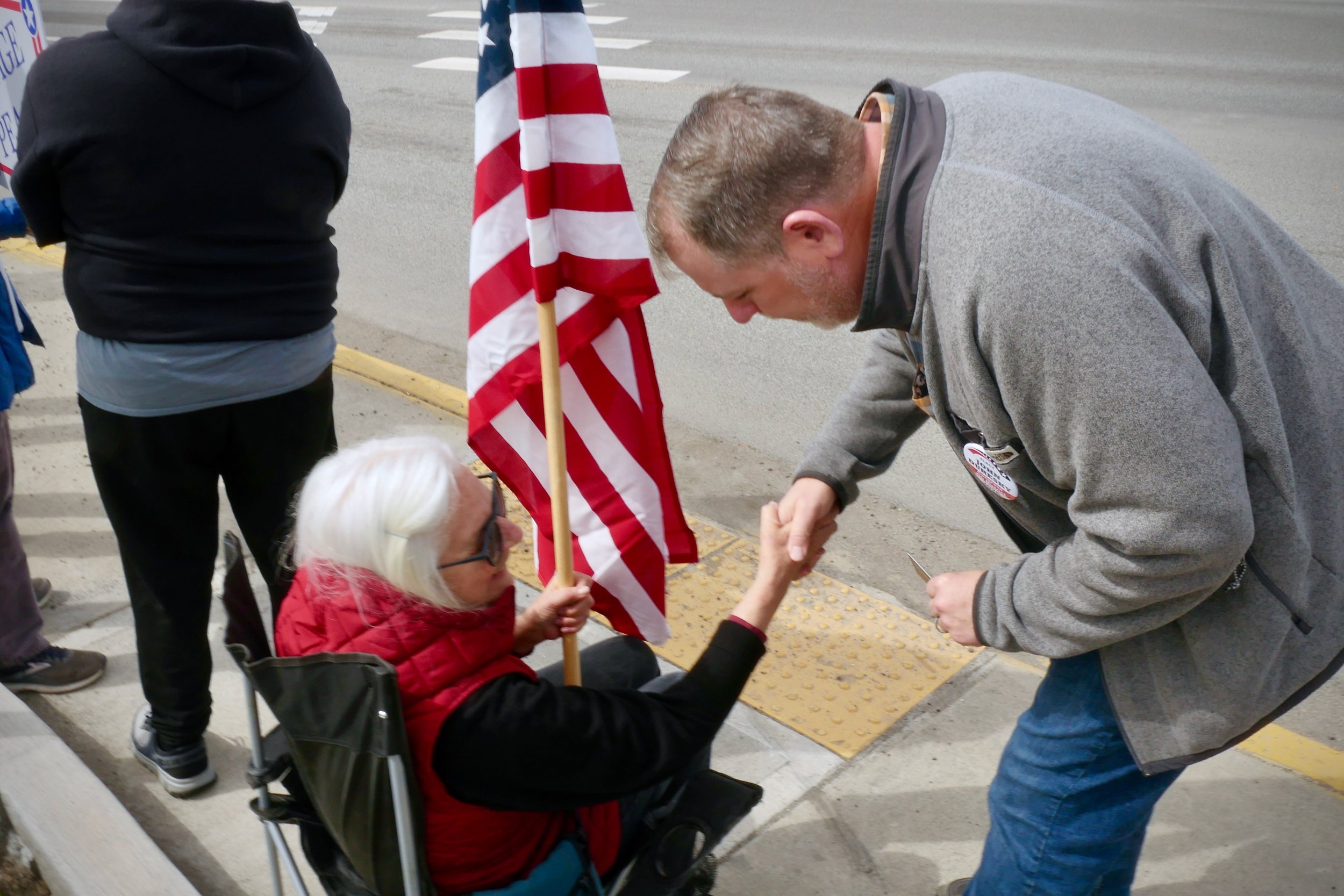 An elderly woman with white hair and glasses, sitting in a wheelchair, holding an American flag, shaking hands with John Duresky on a sidewalk in Omak, WA.