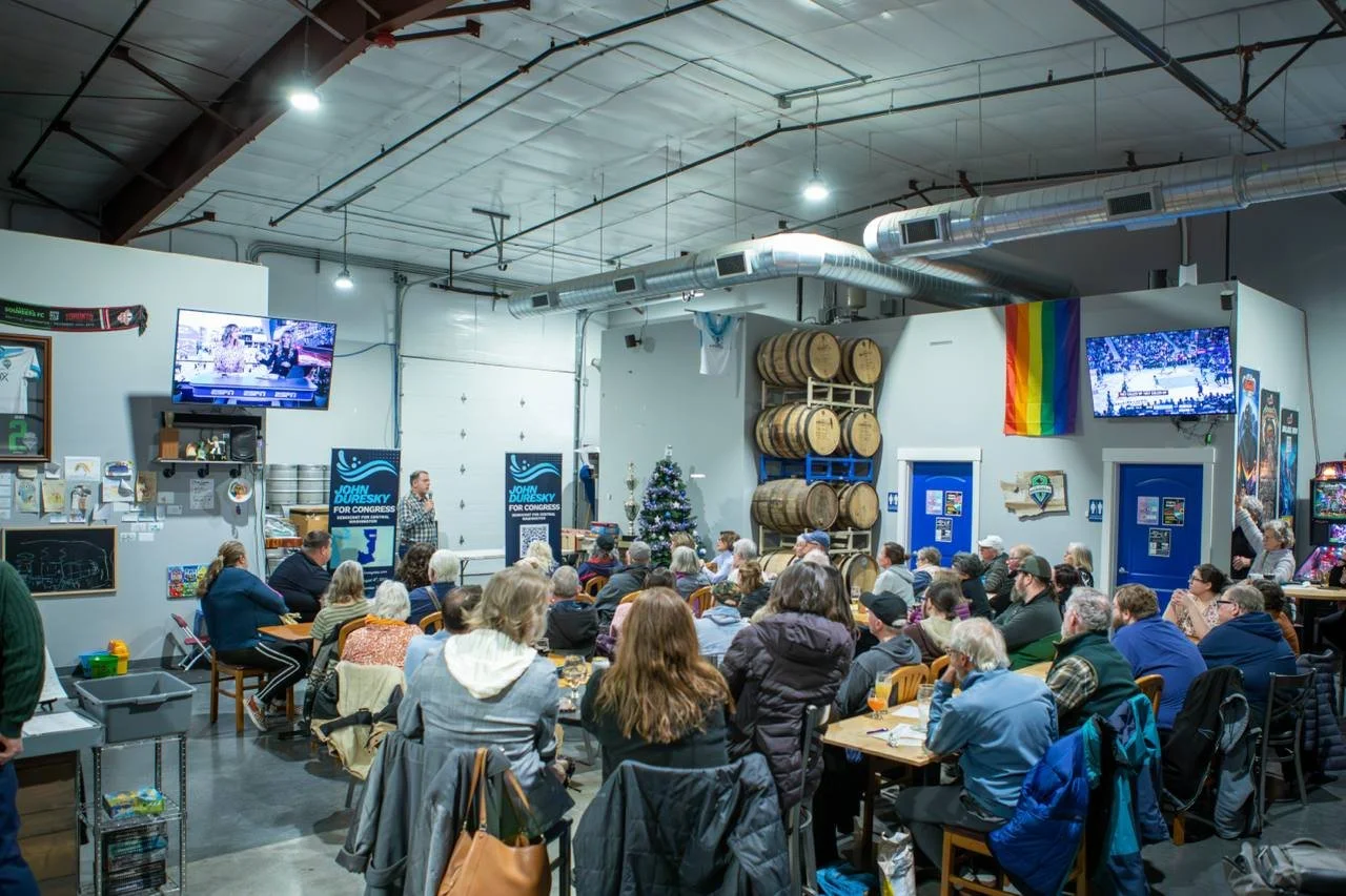 People attending a political event or community gathering in a brewery, with a Christmas tree, a rainbow flag, and large barrels, watching a speaker on stage.