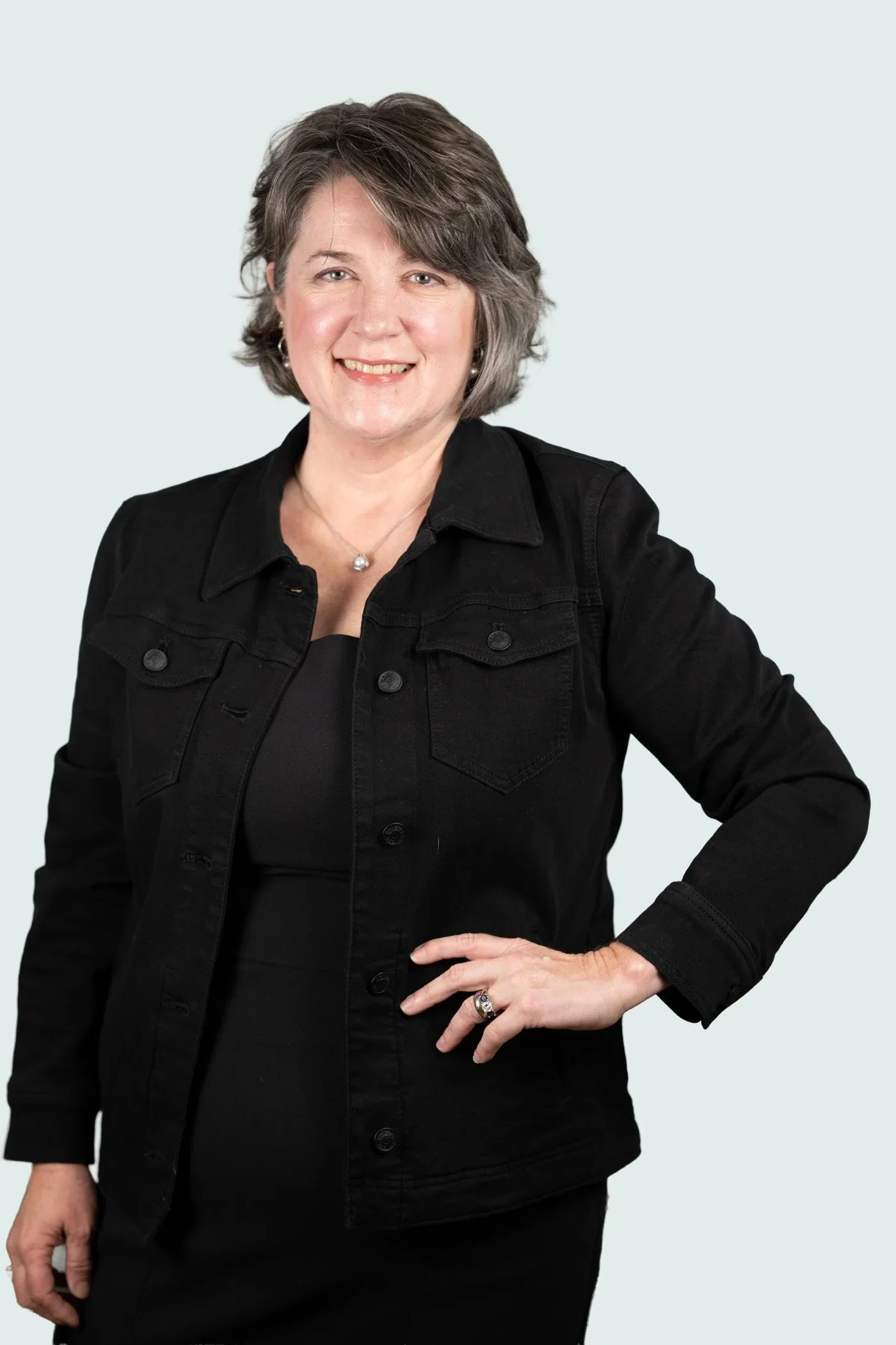 Anne Michael Greene wearing a black shirt and black jacket, smiling with her right hand on her hip, against a pale blue background.