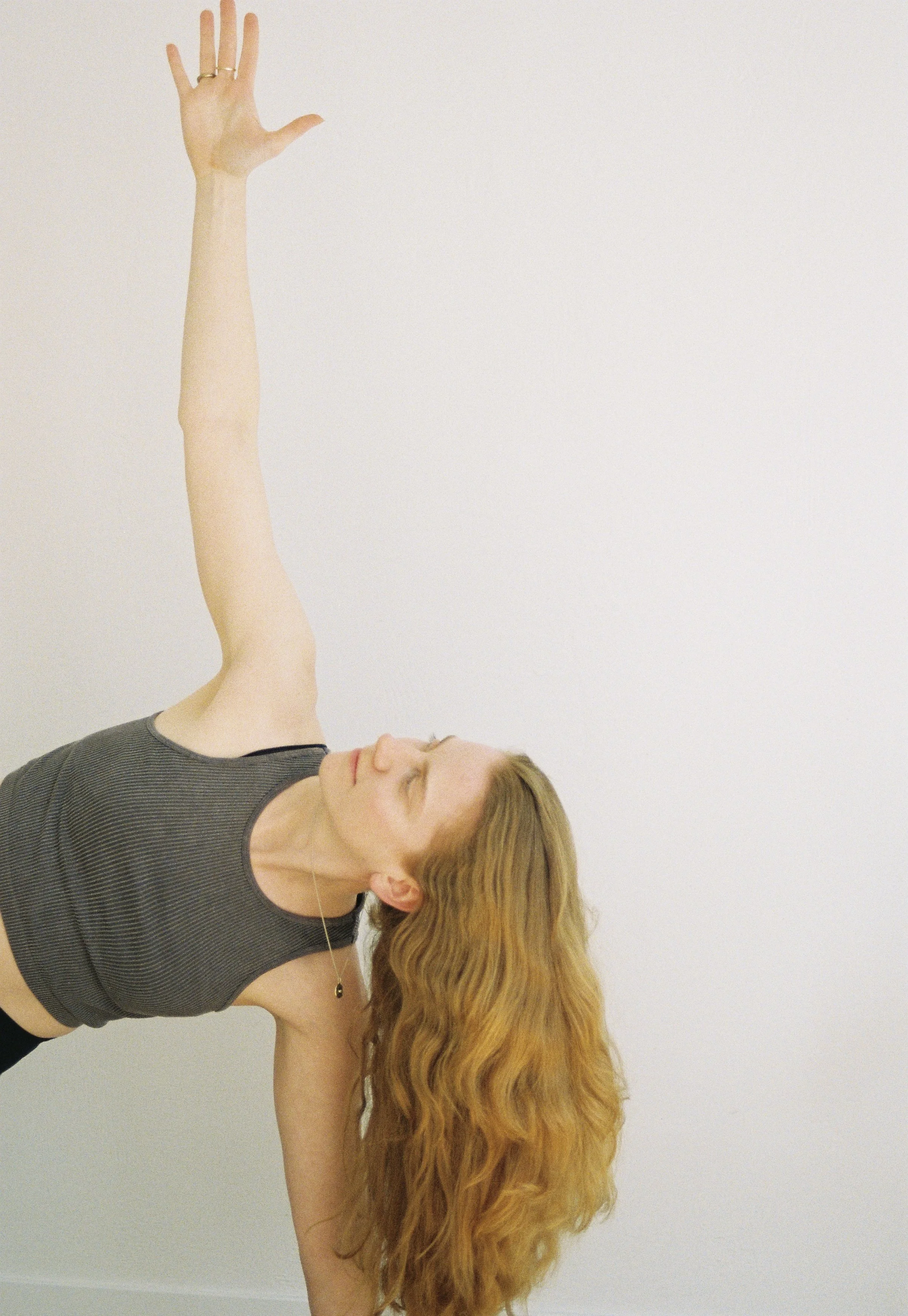 Eine Frau macht eine Yoga-Übung, bei der sie ihren Oberkörper nach hinten beugt und den Arm nach oben streckt, während sie aufrecht steht, vor einem weißen Hintergrund.