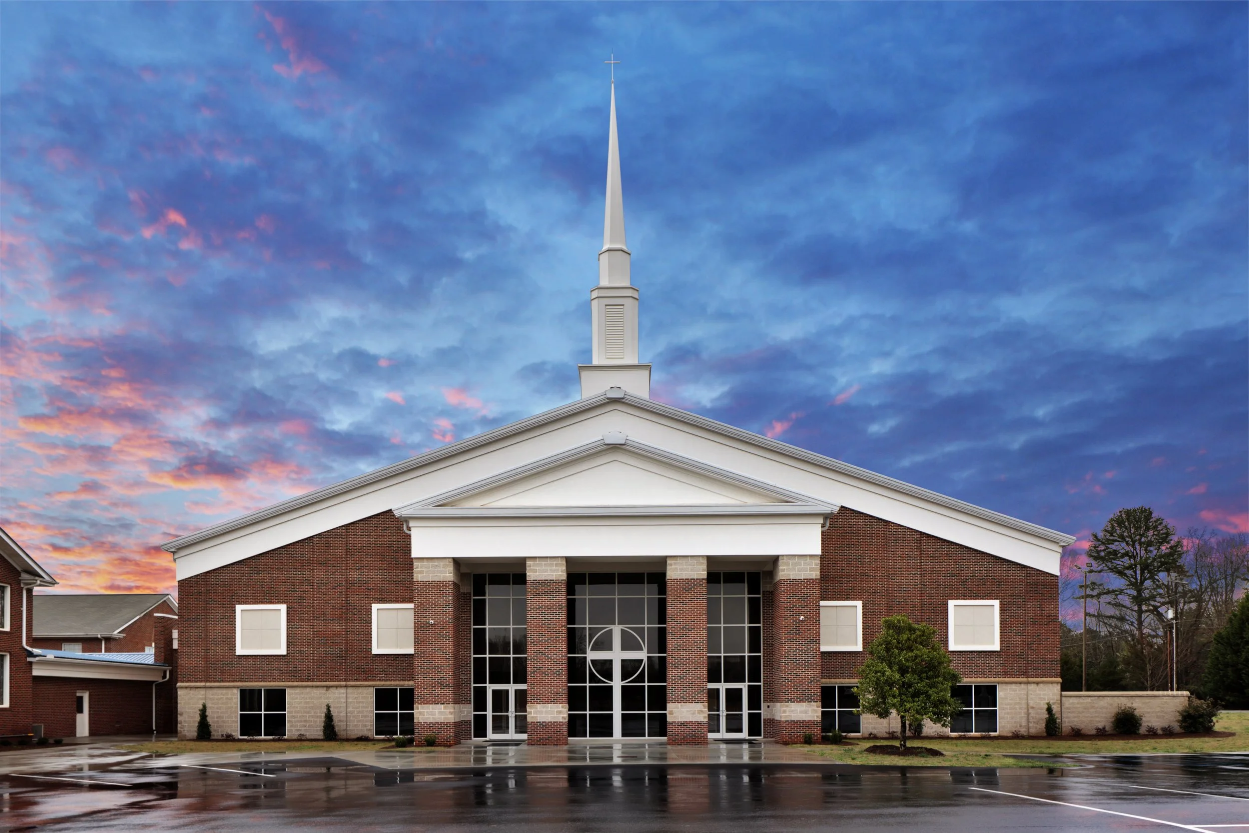 Modern brick church building with tall white steeple and cross, large glass entrance, and dramatic colorful sunset sky reflecting on a wet parking lot.