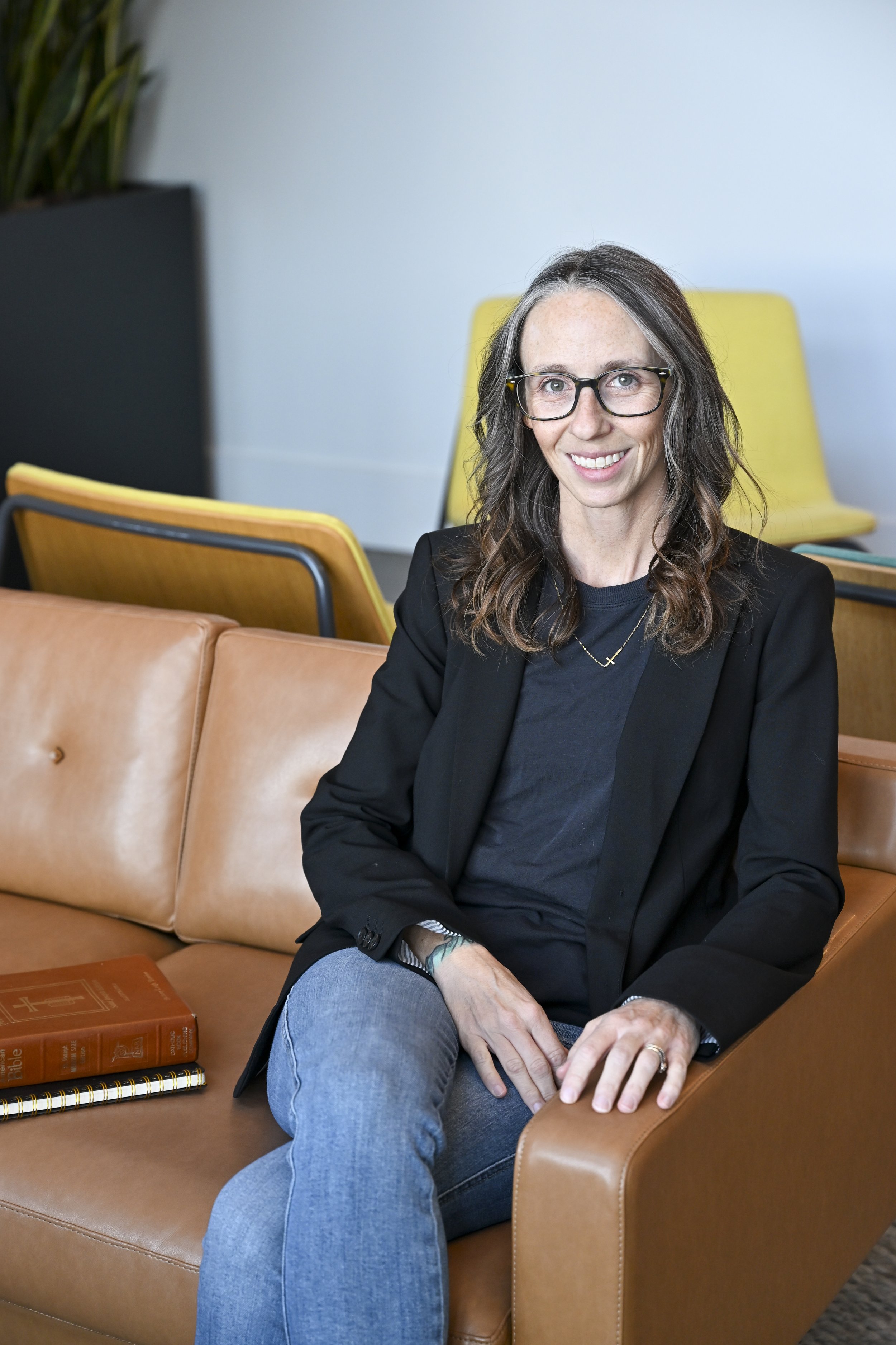 Smiling woman wearing glasses in black blazer, seated on tan sofa in modern office with a bible and notebook beside her.