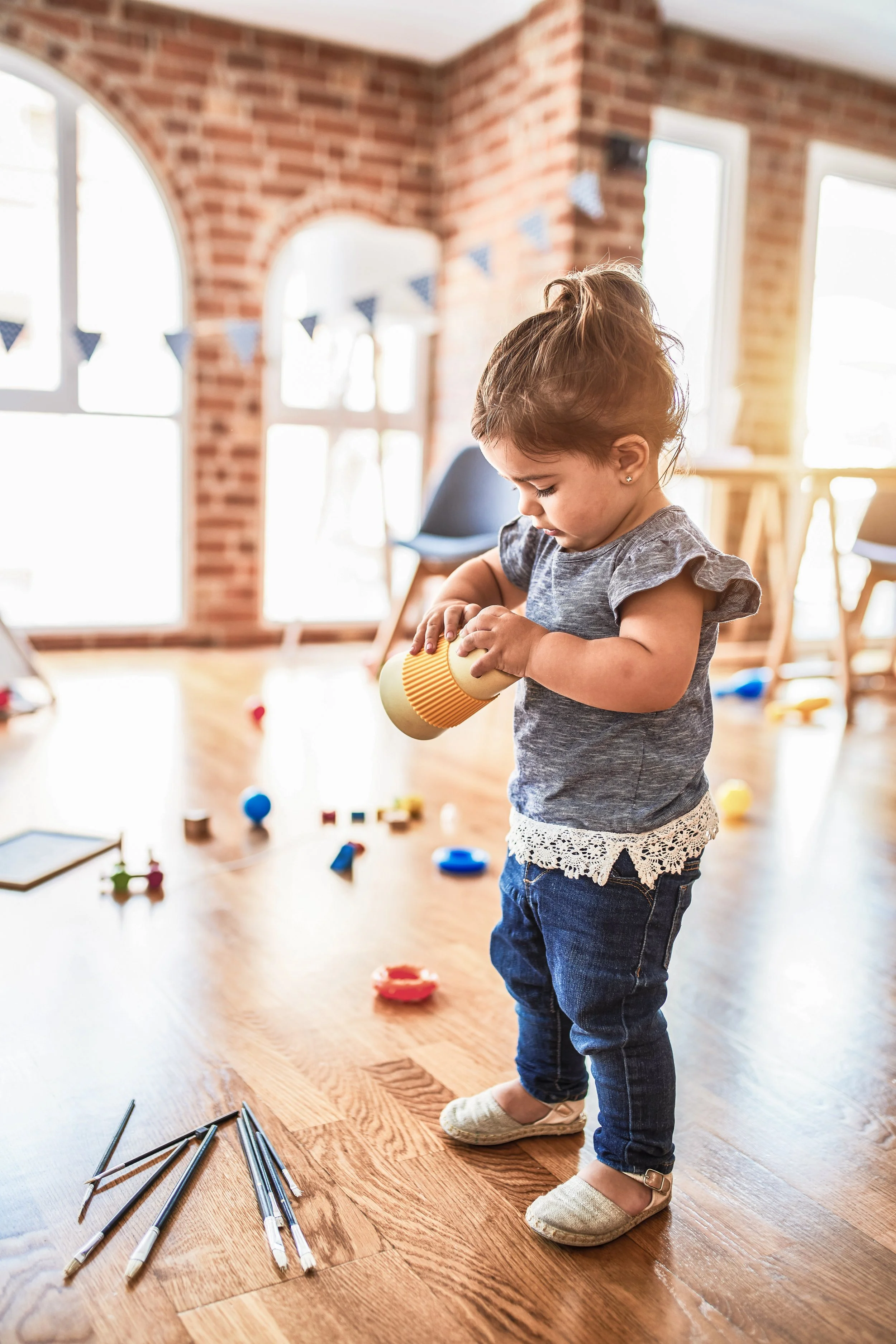 A young girl playing with toys and building blocks on a wooden floor in a sunlit room with brick walls and large windows.