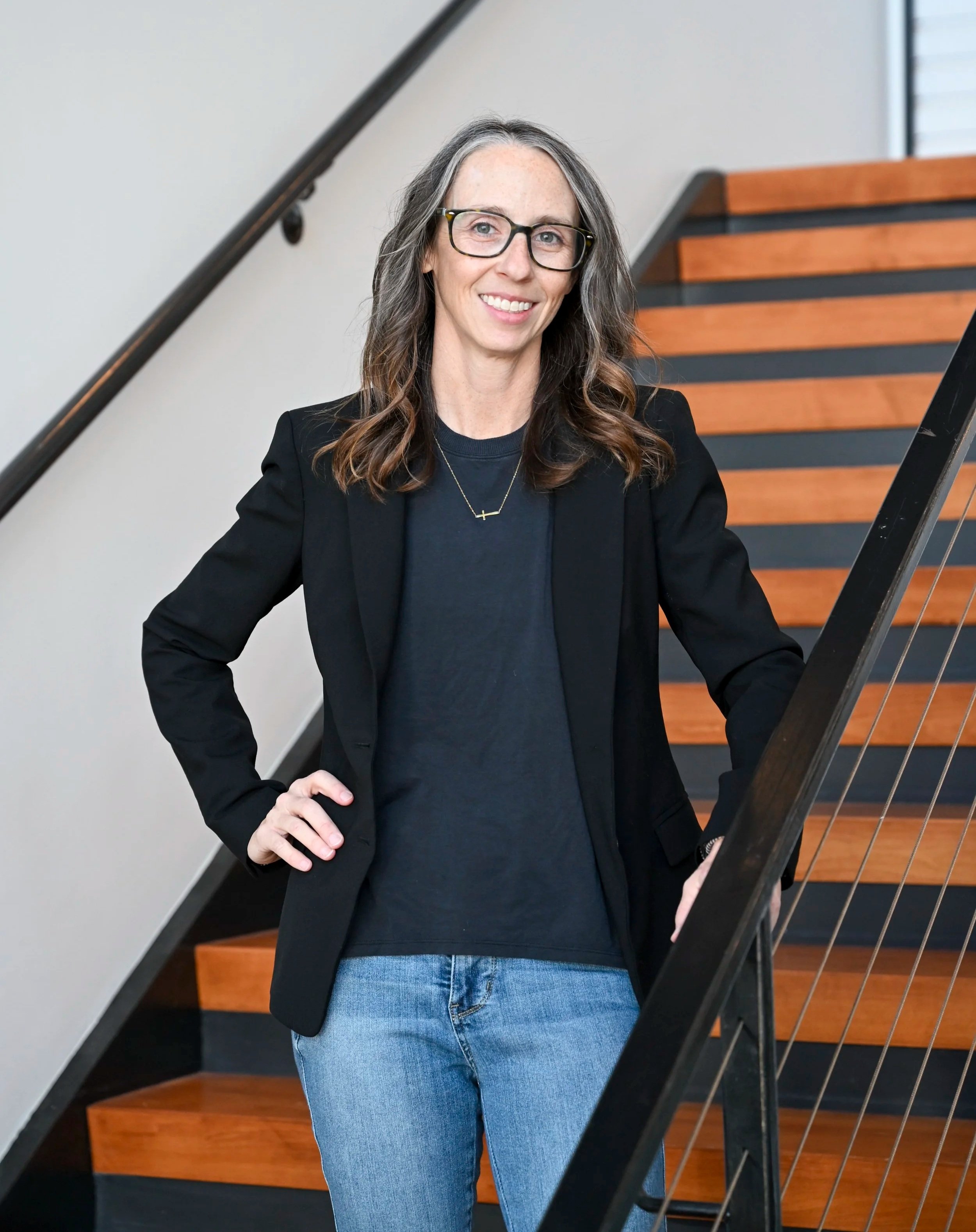 Woman in navy shirt and dark blazer, smiling standing on a staircase, holding a railing.