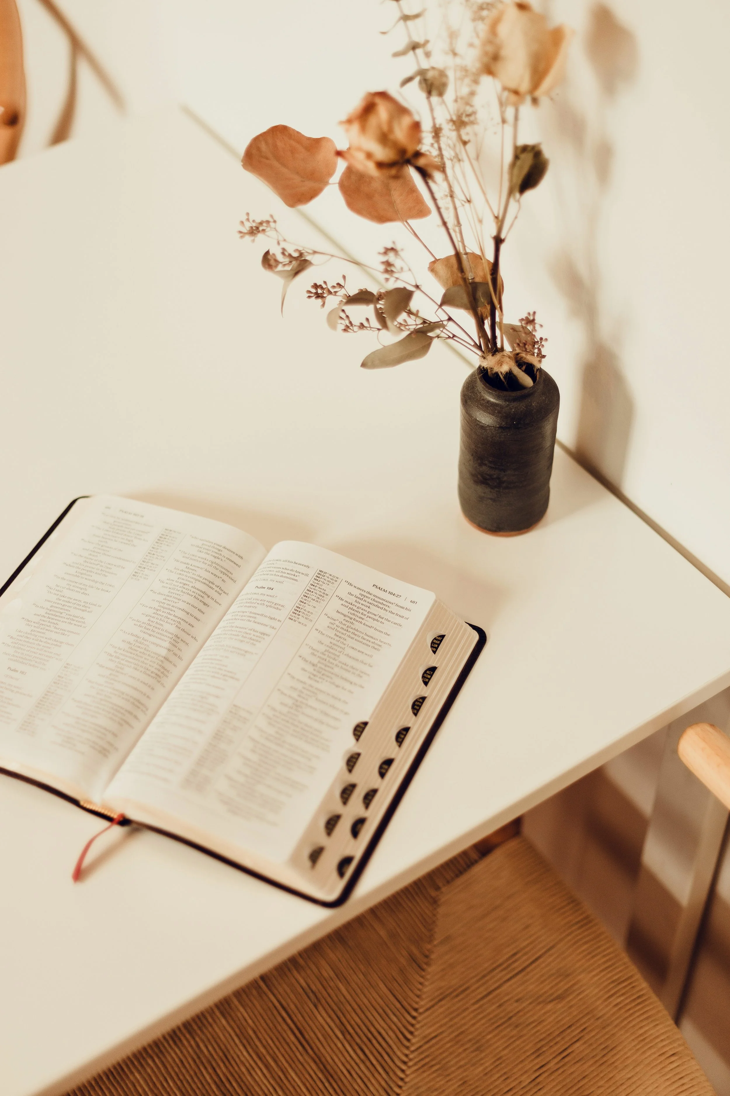 A white table with an open Bible and a black ceramic vase holding dried flowers.