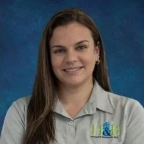A woman with long brown hair wearing a light gray collared shirt and small earrings, smiling against a blue background.