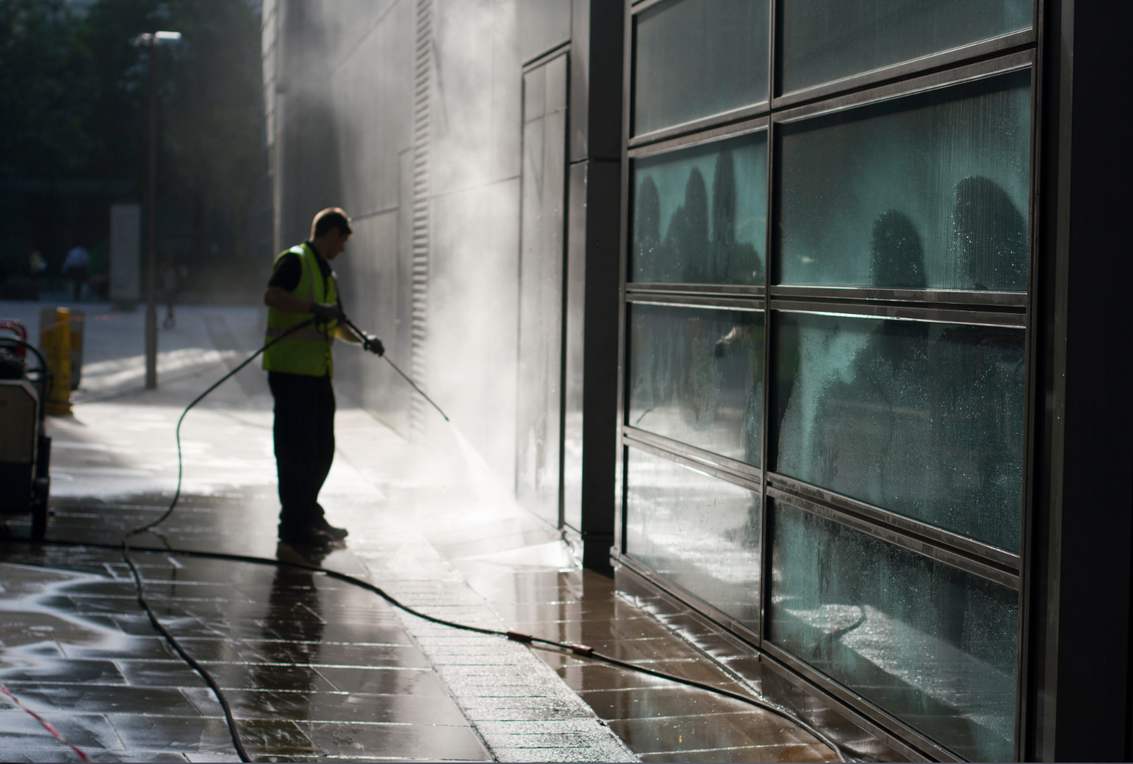 A worker in a yellow safety vest pressure washing the sidewalk outside a modern building with glass windows.