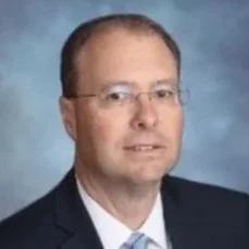 Professional headshot of a man wearing glasses, a dark suit, and a light-colored tie, with a blue background