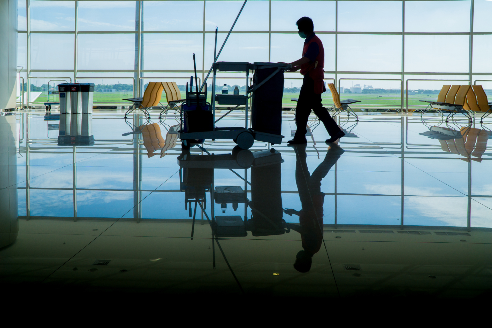 Silhouette of an airport janitorial service worker pushing a cart with cleaning supplies in front of large windows showing a runway and blue sky outside.