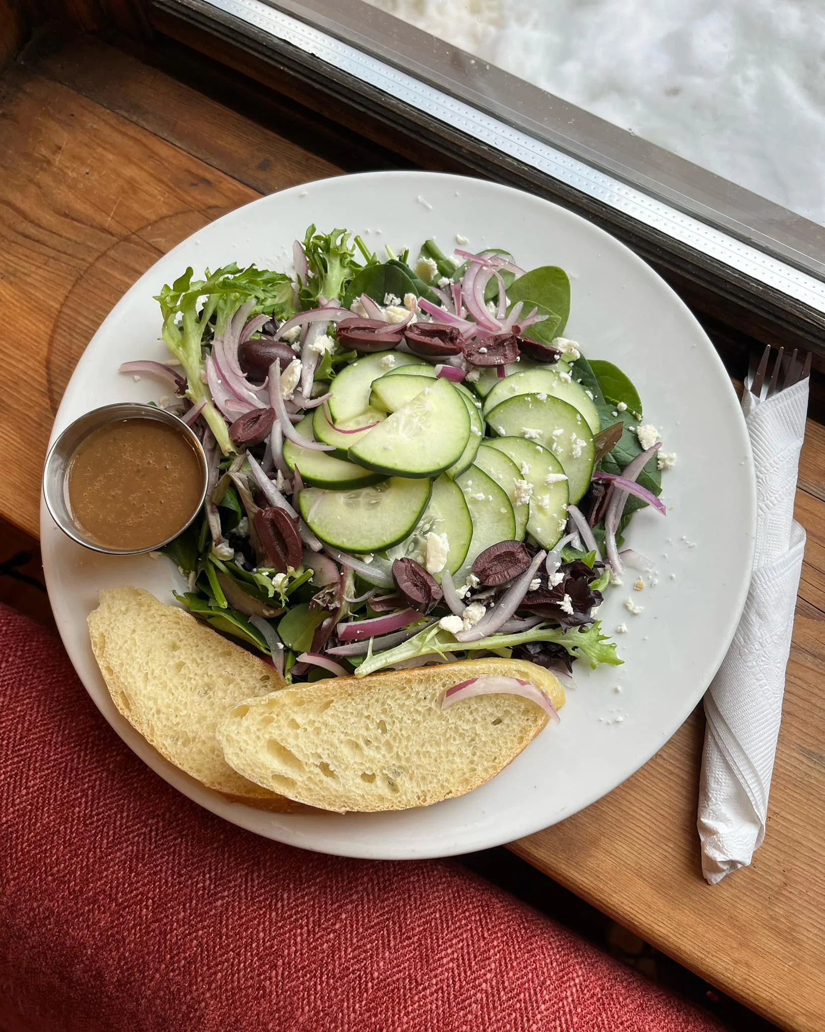 A white plate with a mixed green salad containing cucumber slices, red onion slices, black olives, and crumbled feta cheese, served with two slices of bread and a small container of salad dressing, placed on a wooden table near a window.