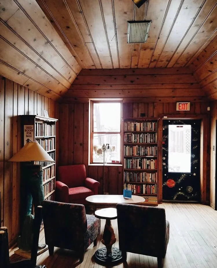 Bookshelves filled with books, a red armchair, a small round table, two dark patterned chairs, a floor lamp, a window, and a door decorated with space-themed stickers inside a wooden-paneled room.