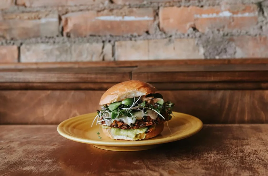 A burger with a bun, green sprouts, melted cheese, a veggie patty, and avocado slices on a yellow plate against a brick wall background.