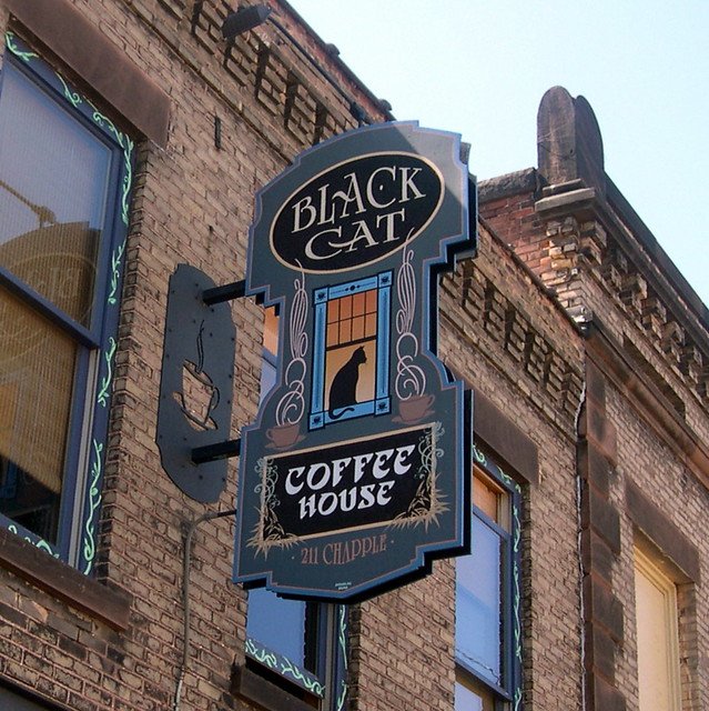 Sign for Black Cat Coffeehouse with a black cat silhouette, brick building exterior, and decorative design at 211 Chapple Avenue.