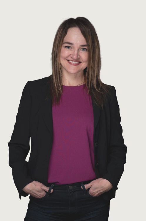 A woman with shoulder-length brown hair, wearing a black blazer over a purple top, smiling and posing with her hands in her pockets against a plain white background.