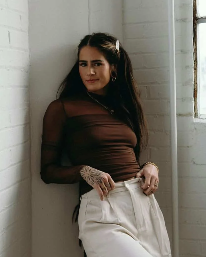 Young woman with dark hair and hoop earrings leaning against a white brick wall, wearing a sheer black top and white high-waisted pants, with tattoos on her hand, in a well-lit room with a small window.