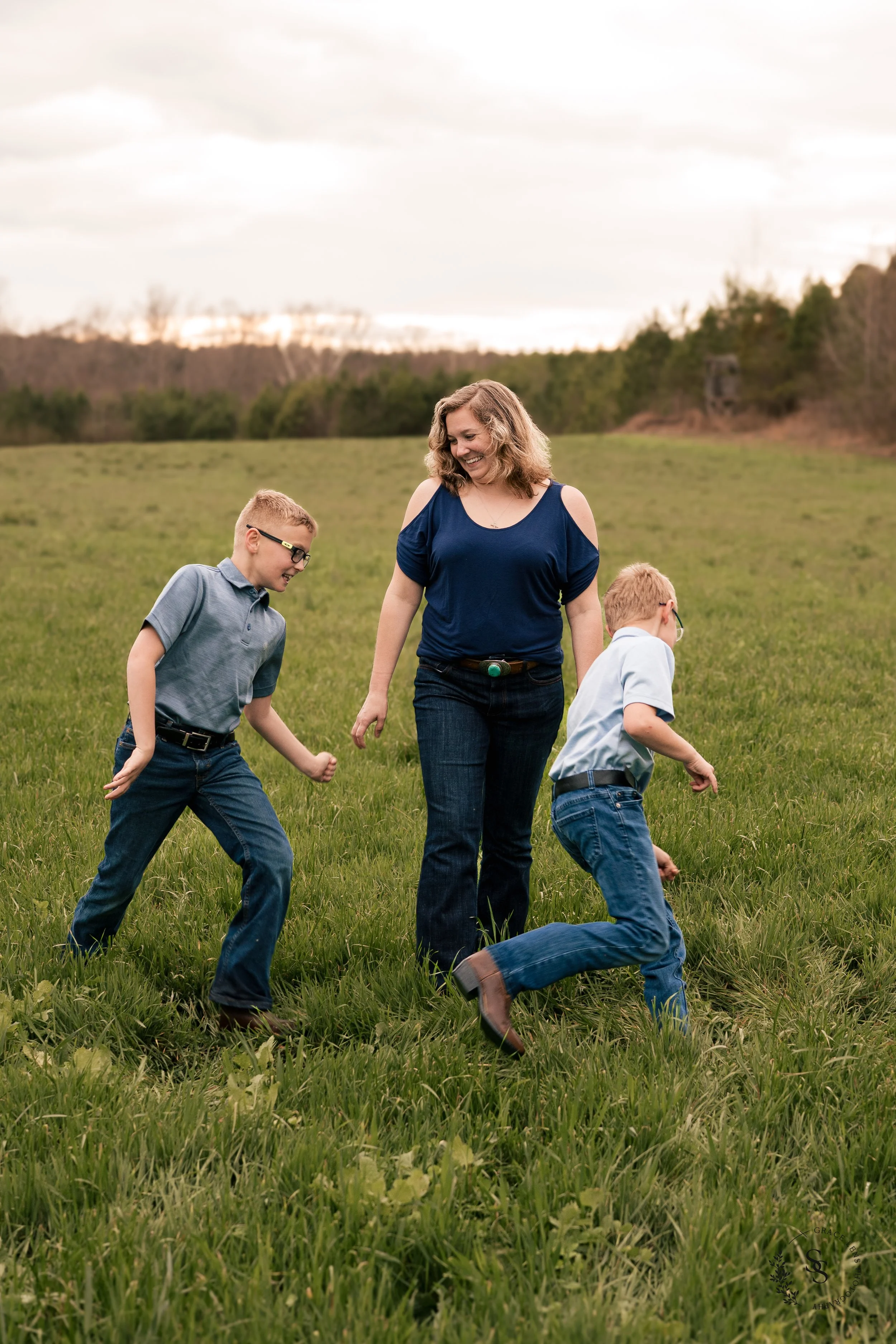 A woman and two young boys playing and running on a grassy field outdoors during daytime, with a cloudy sky and trees in the background.