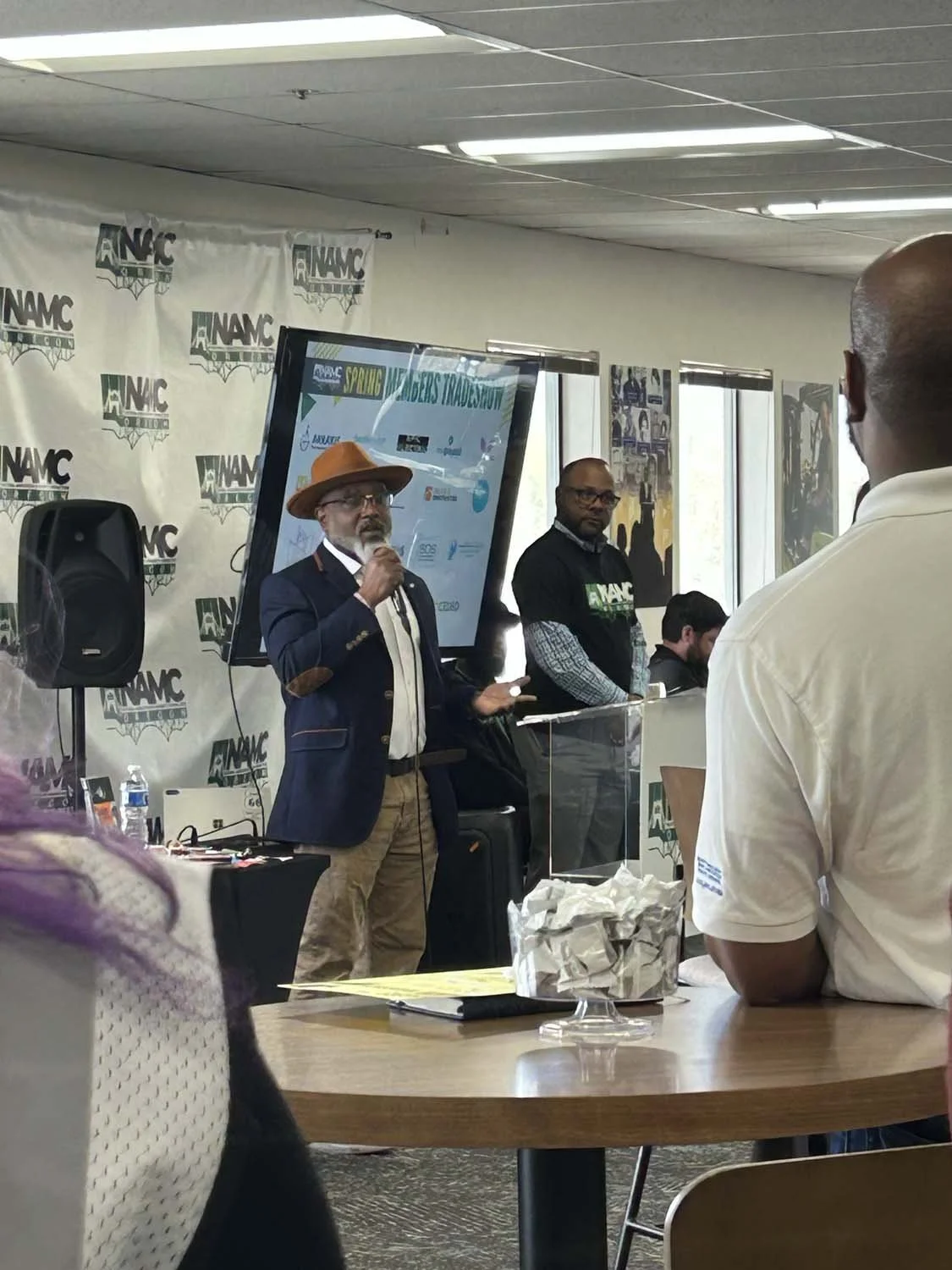 Herman Greene with glasses, a beard, and a brown hat speaking into a microphone at a conference with people seated around. There is a banner with 'NAMC' logo in the background and a large screen displaying the event's title.