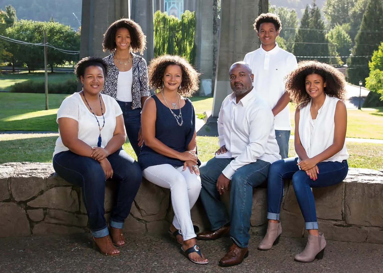 Herman's family of seven, including five women and two men, gathered outdoors in a park with greenery and trees in the background. They are sitting and standing on and beside a stone wall, smiling at the camera on a sunny day.