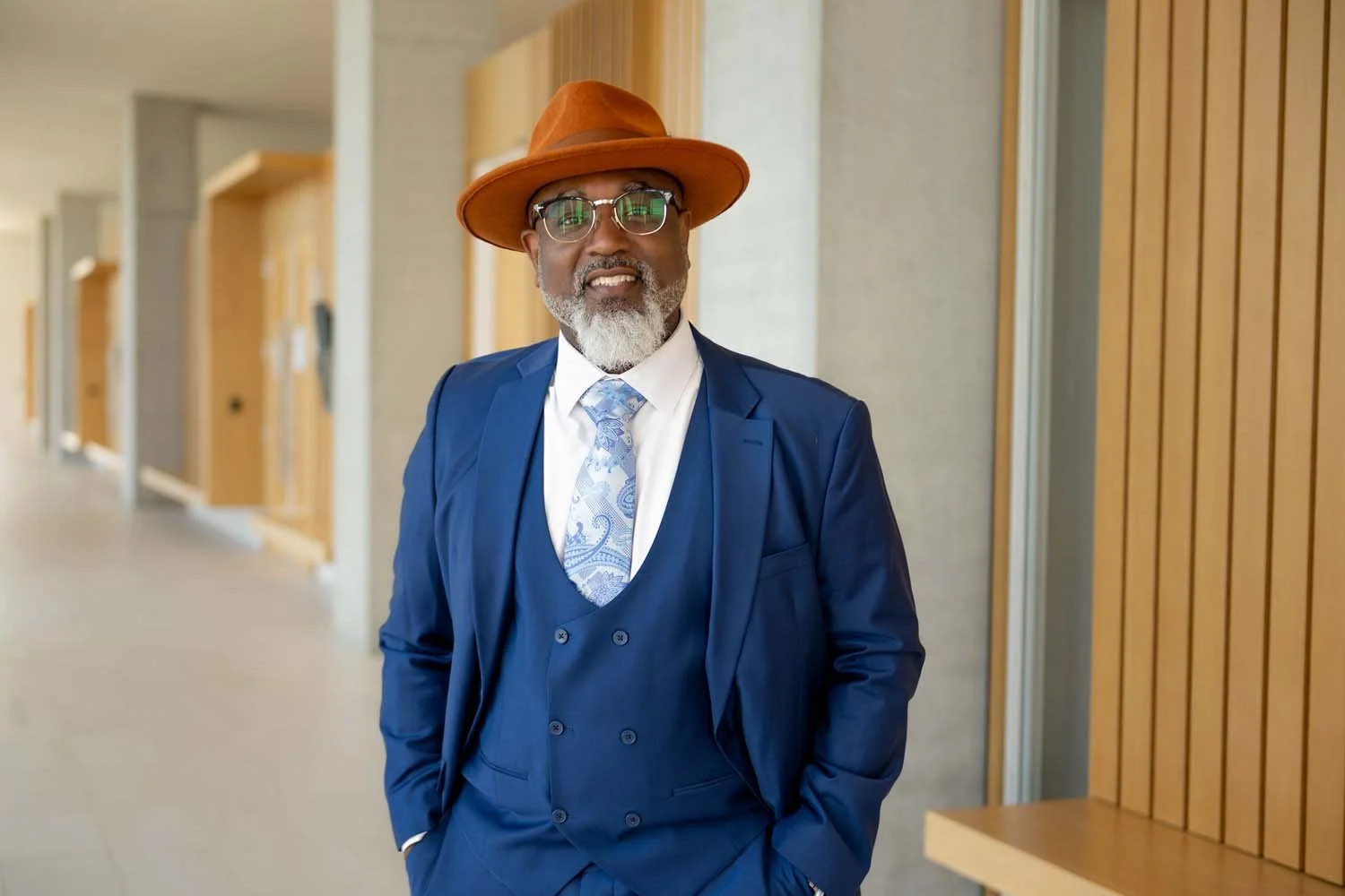 Herman Greene running for Multnomah County Commissioner District 2, shown here in a blue suit, white shirt, and patterned tie, wearing glasses and a brown hat.