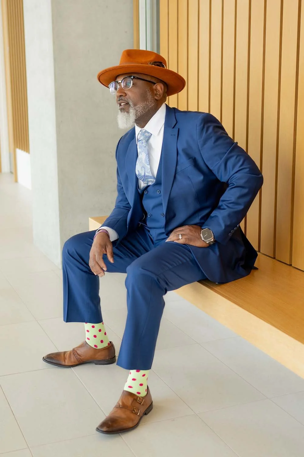 Herman Greene in a blue suit with a brown hat, polka-dot socks, and brown shoes, sitting on a bench against a wooden-paneled wall.