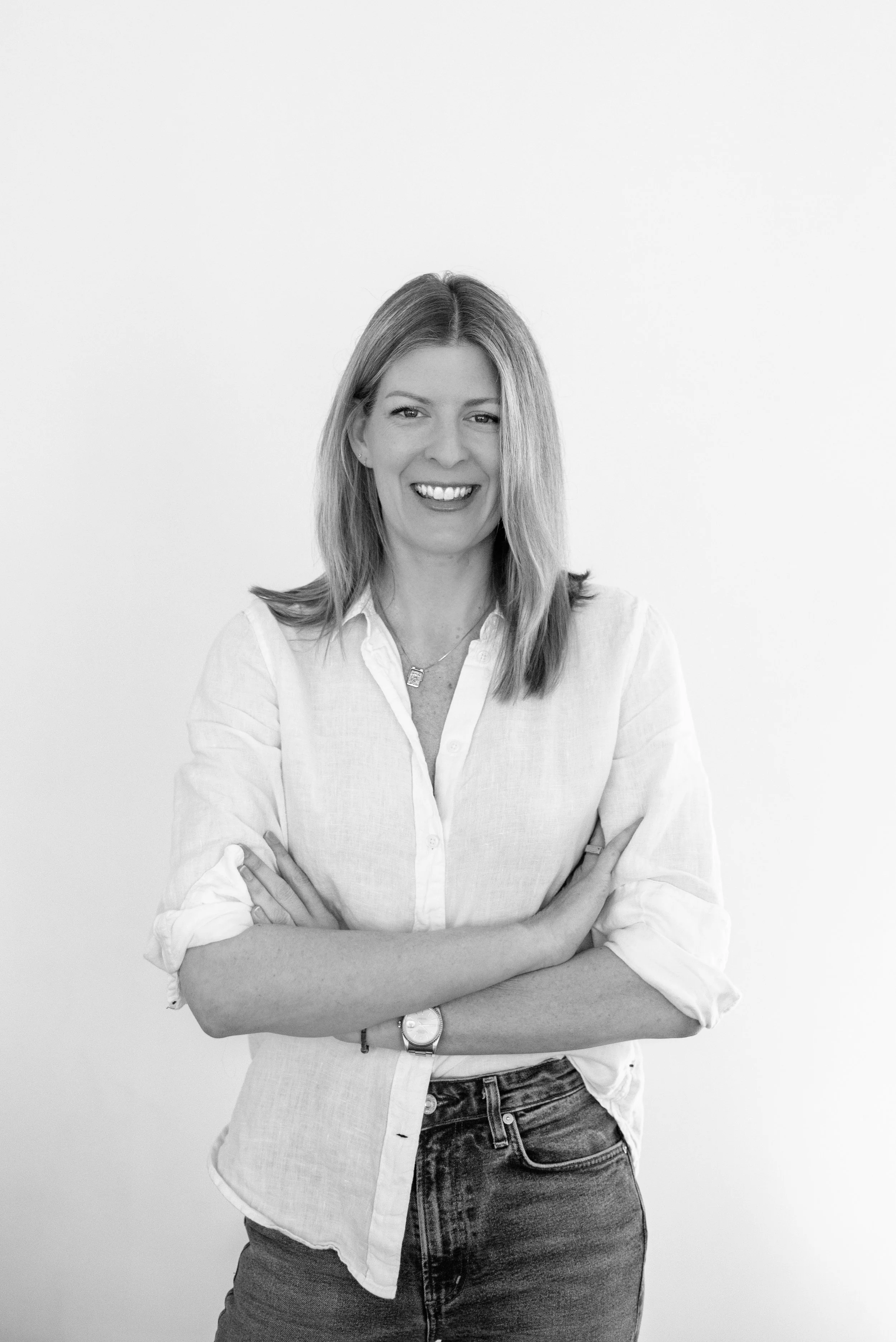 Black and white photo of a smiling woman with long hair, wearing a white button-up shirt and jeans, crossing her arms, against a plain background.