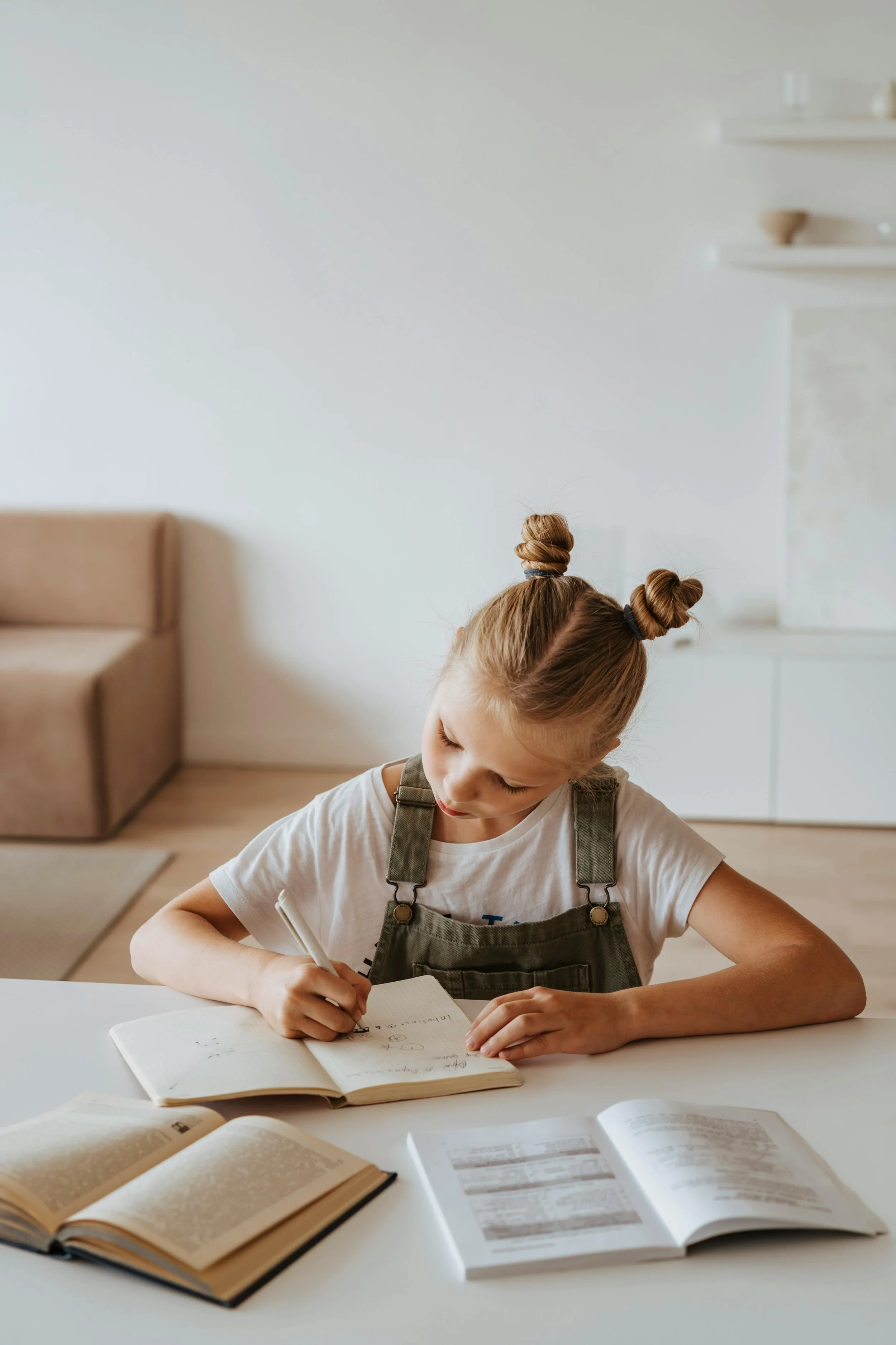A young girl with two buns in her hair is writing in a notebook at a white table, surrounded by open books, in a bright room.