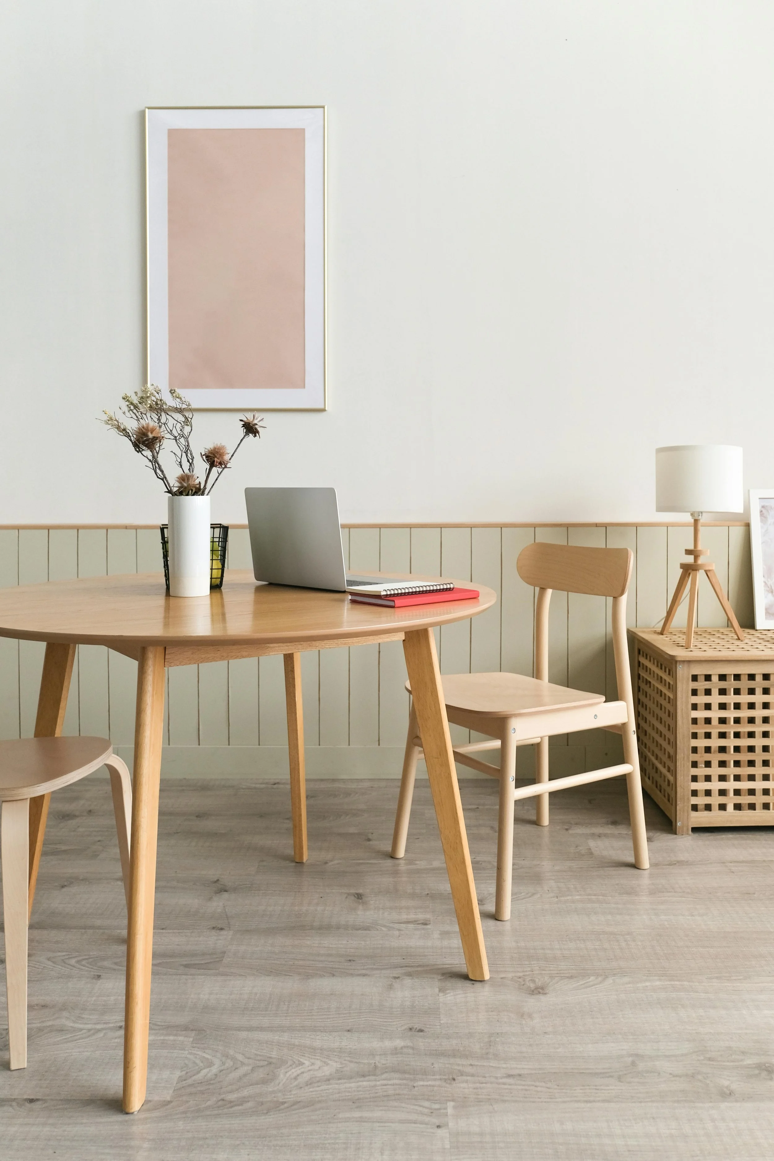 A minimalist room with a light-colored wooden table, a laptop, a red notebook, and a vase with dried flowers, against a cream wall with artwork and a small side table with a lamp.