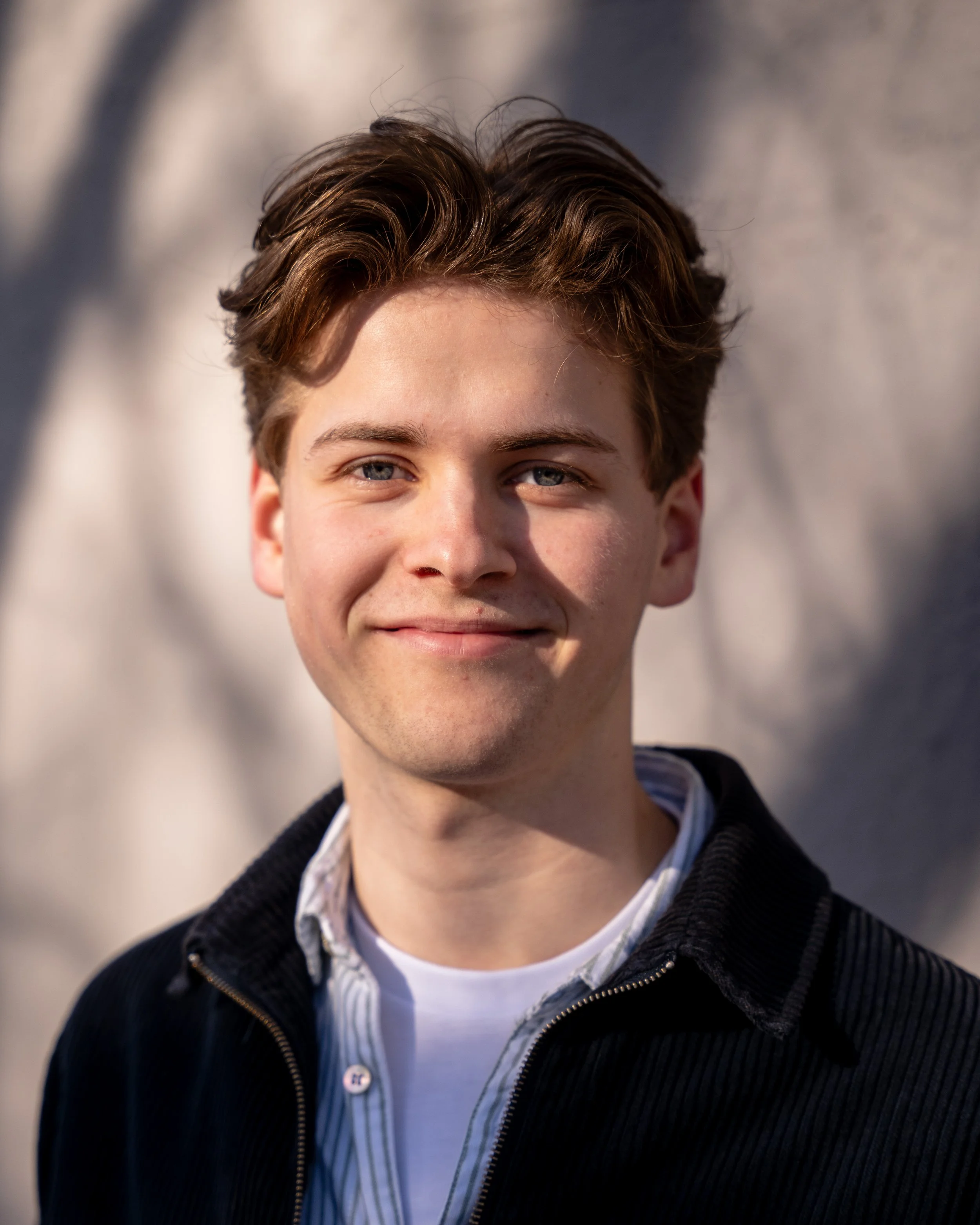 Close-up of a young man with brown hair, blue eyes, and fair skin smiling outdoors with a blurred background.