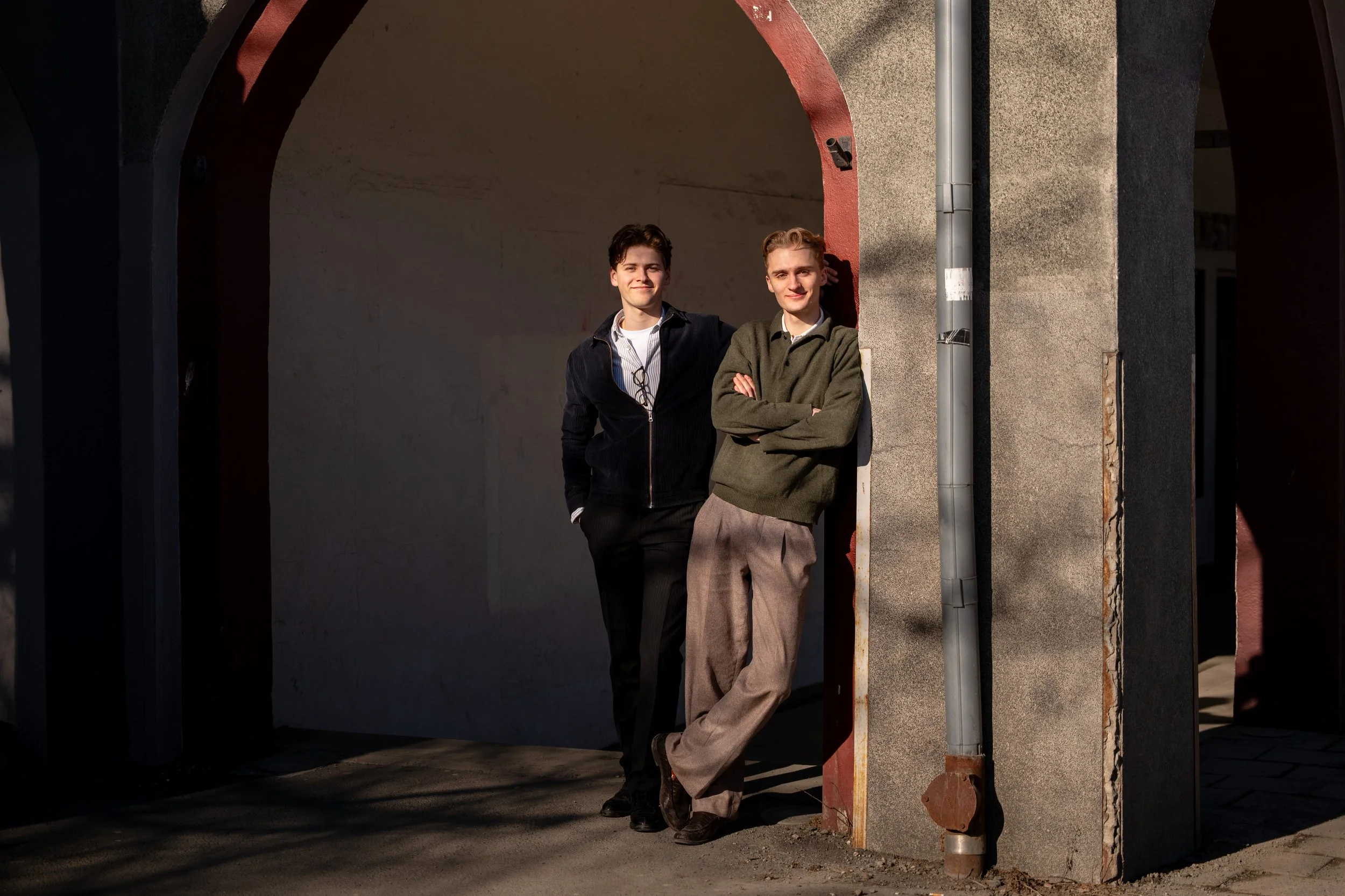 To unge menn står under en buegang ved en vegg og smiler mot kamera. Den ene har på svart jakke og mørke bukser, den andre grønn genser og beige bukser.