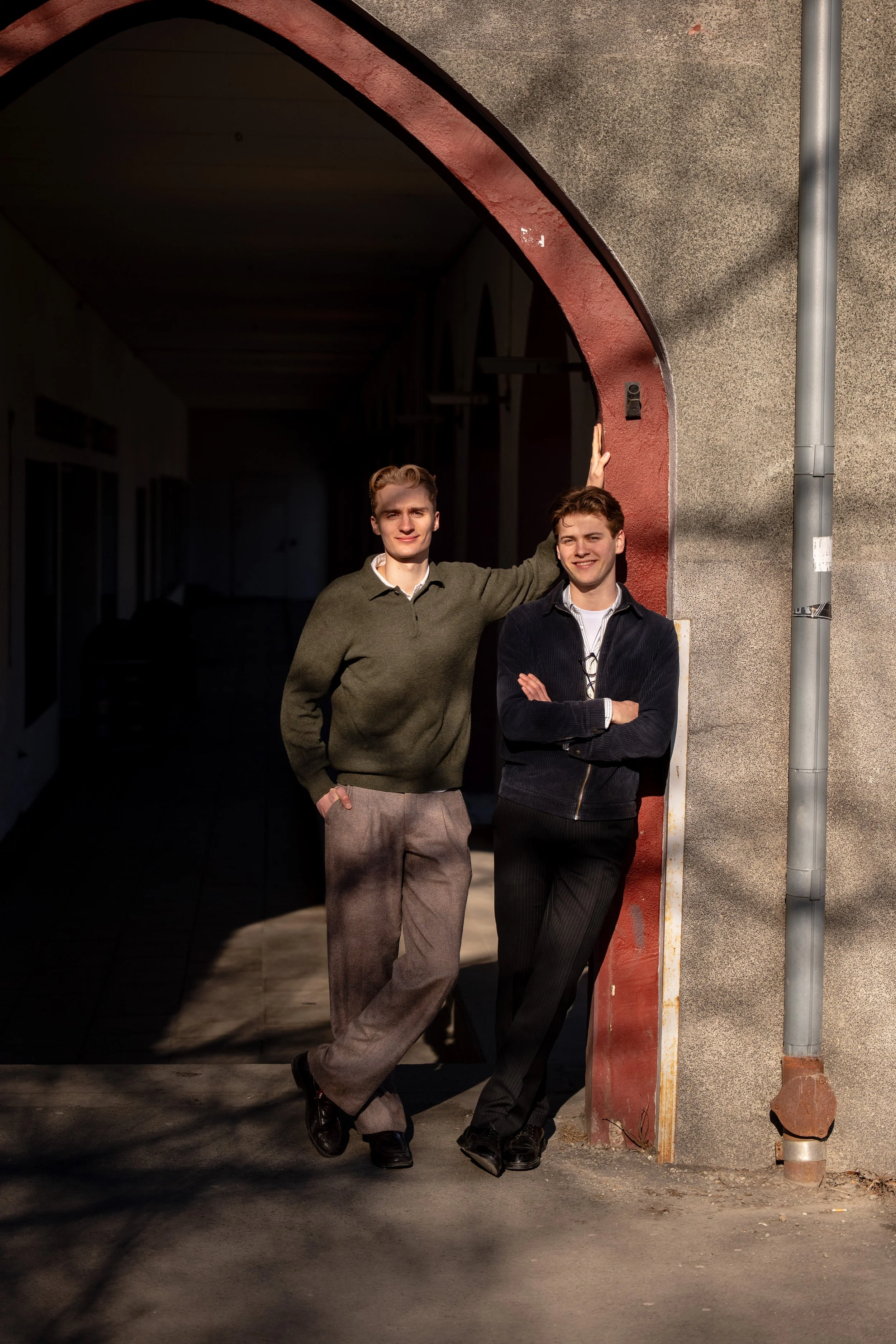 Two young men standing by the entrance of a building with an arched doorway, one with his hand raised against the wall, both smiling and dressed casually.