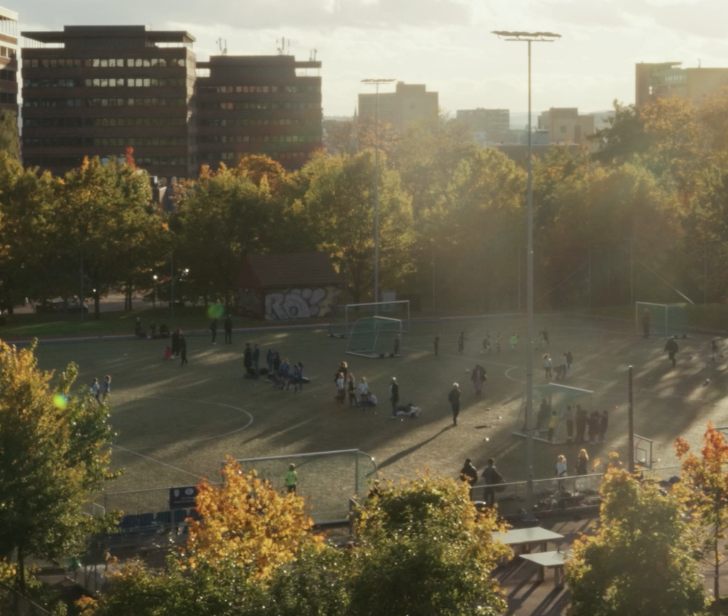A city park in autumn with trees showing fall colors, people playing soccer and sitting on benches, and tall buildings in the background. The sunlight creates a warm glow.