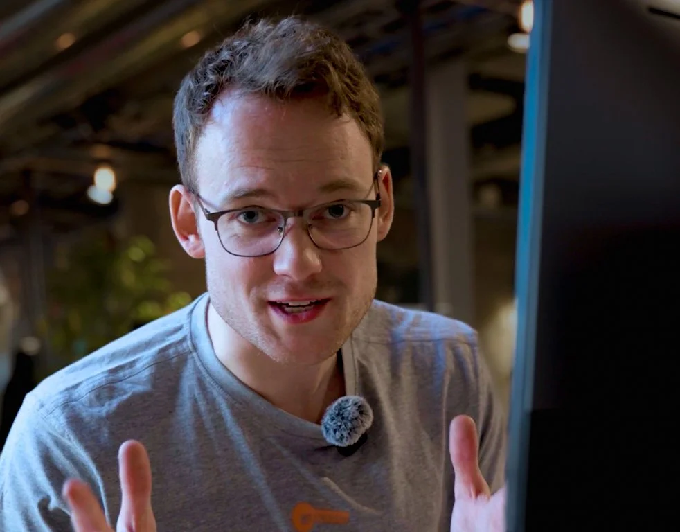 A man with glasses wearing a gray shirt, speaking to the camera with a close-up of his face, in a space with a rustic ceiling and dim lighting.