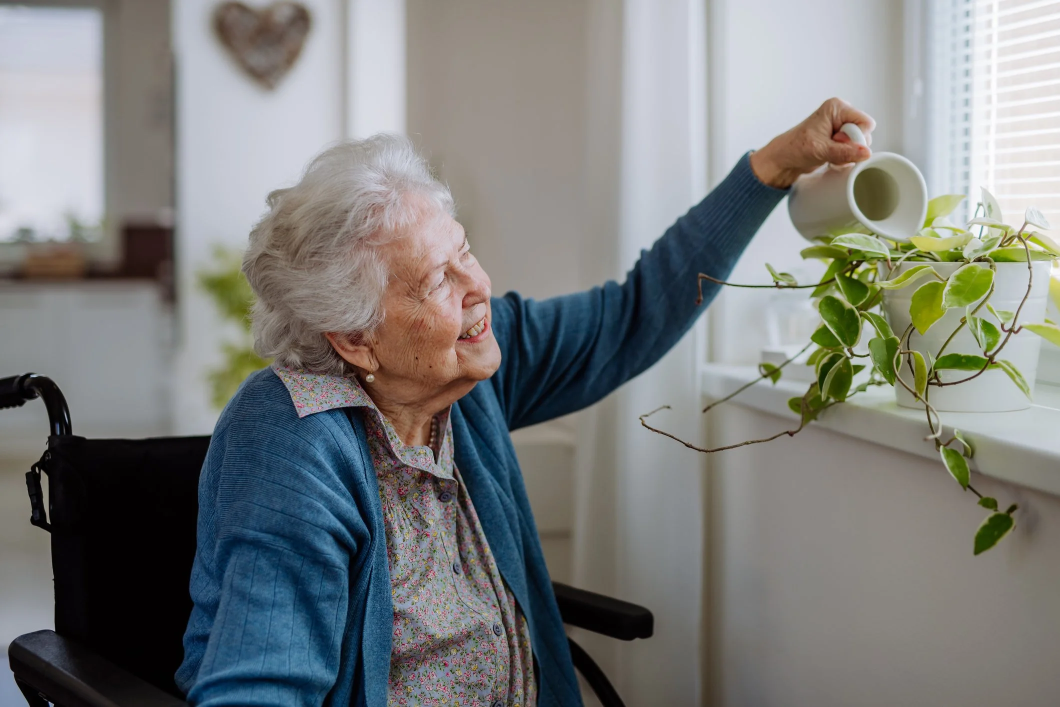 An elderly woman in a wheelchair watering houseplants on a windowsill, smiling.